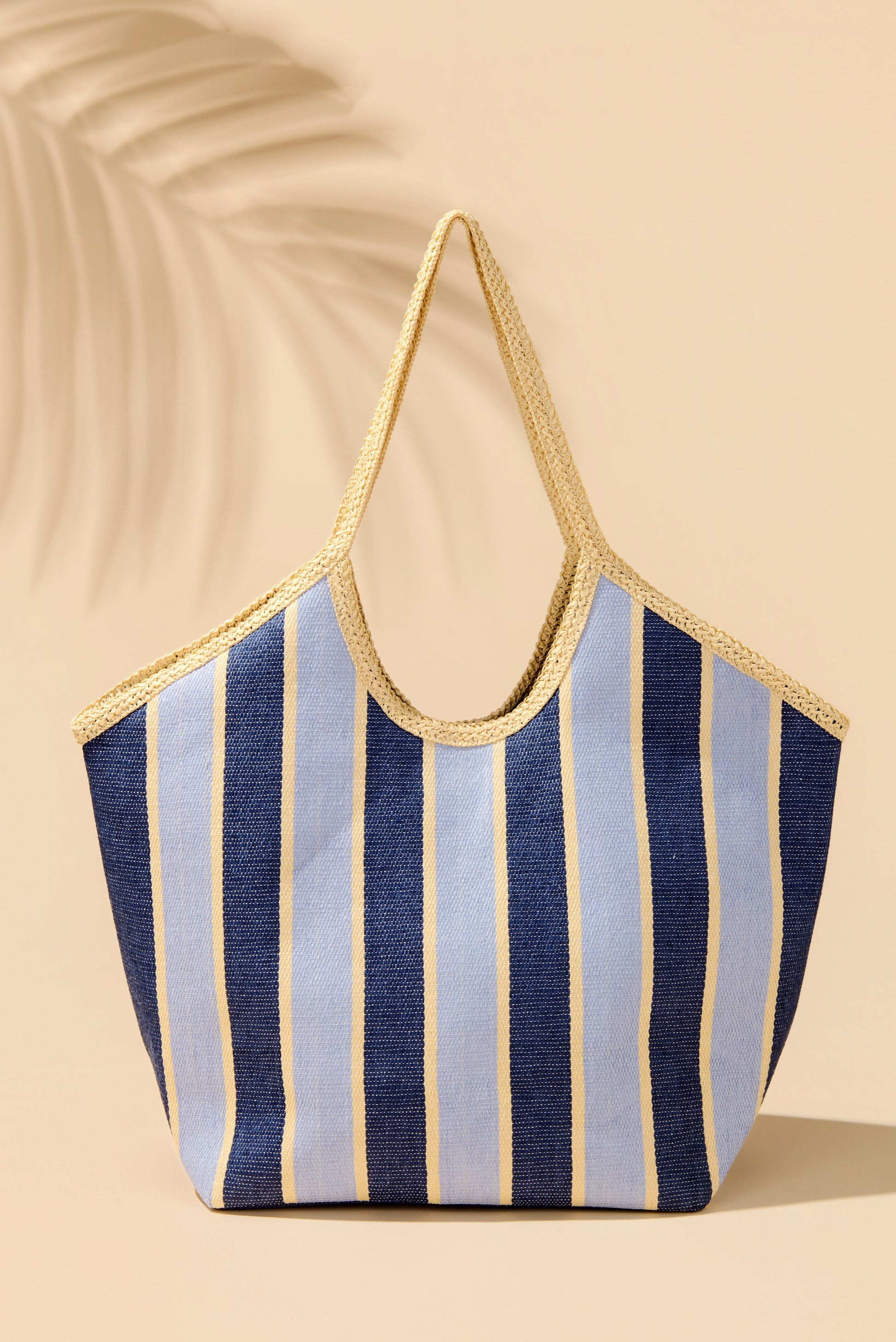 The Capri Tote by South Moon Under is a large woven beach bag in Blue Multi, featuring vertical stripes and beige handles, shown on a beige surface with a palm leaf shadow in the background.