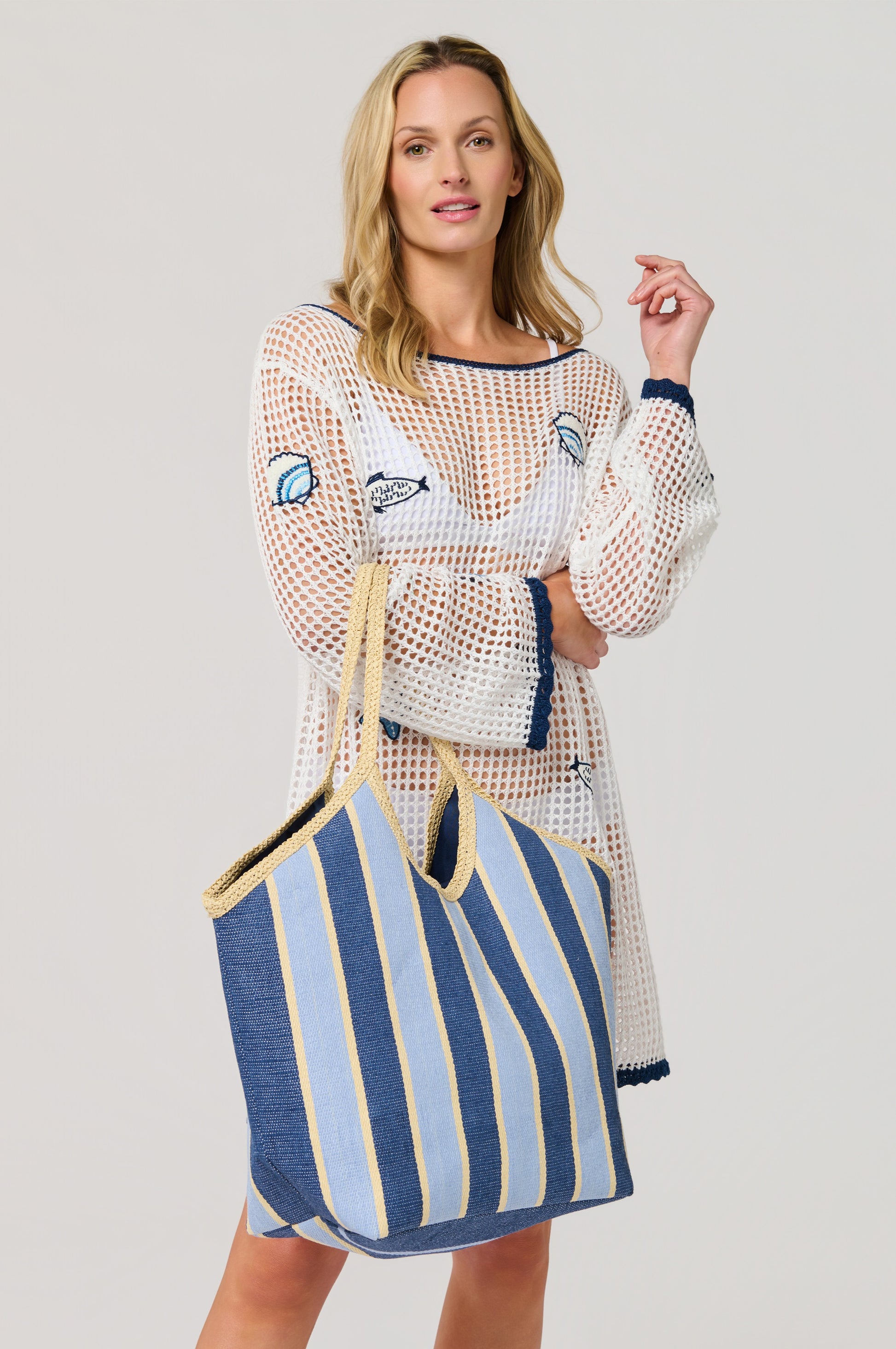 A woman in a white crochet beach cover-up with blue trim and marine motifs holds the South Moon Under Capri Tote, a large blue and beige striped woven straw bag, standing against a plain light background and looking at the camera.
