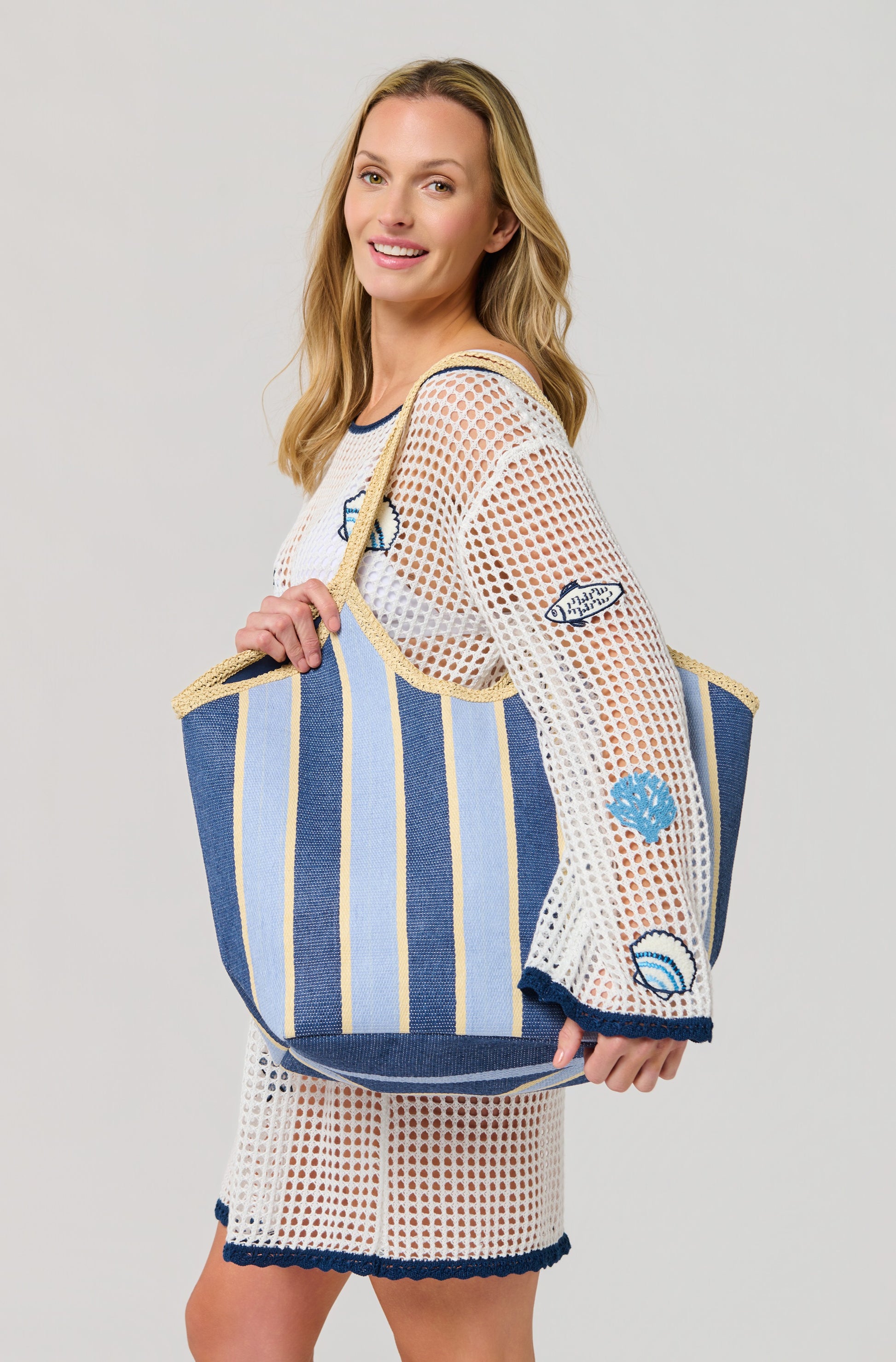 A smiling woman in a white mesh beach cover-up with embroidered seashell and fish details carries the South Moon Under Capri Tote over her shoulder, standing against a plain light background.