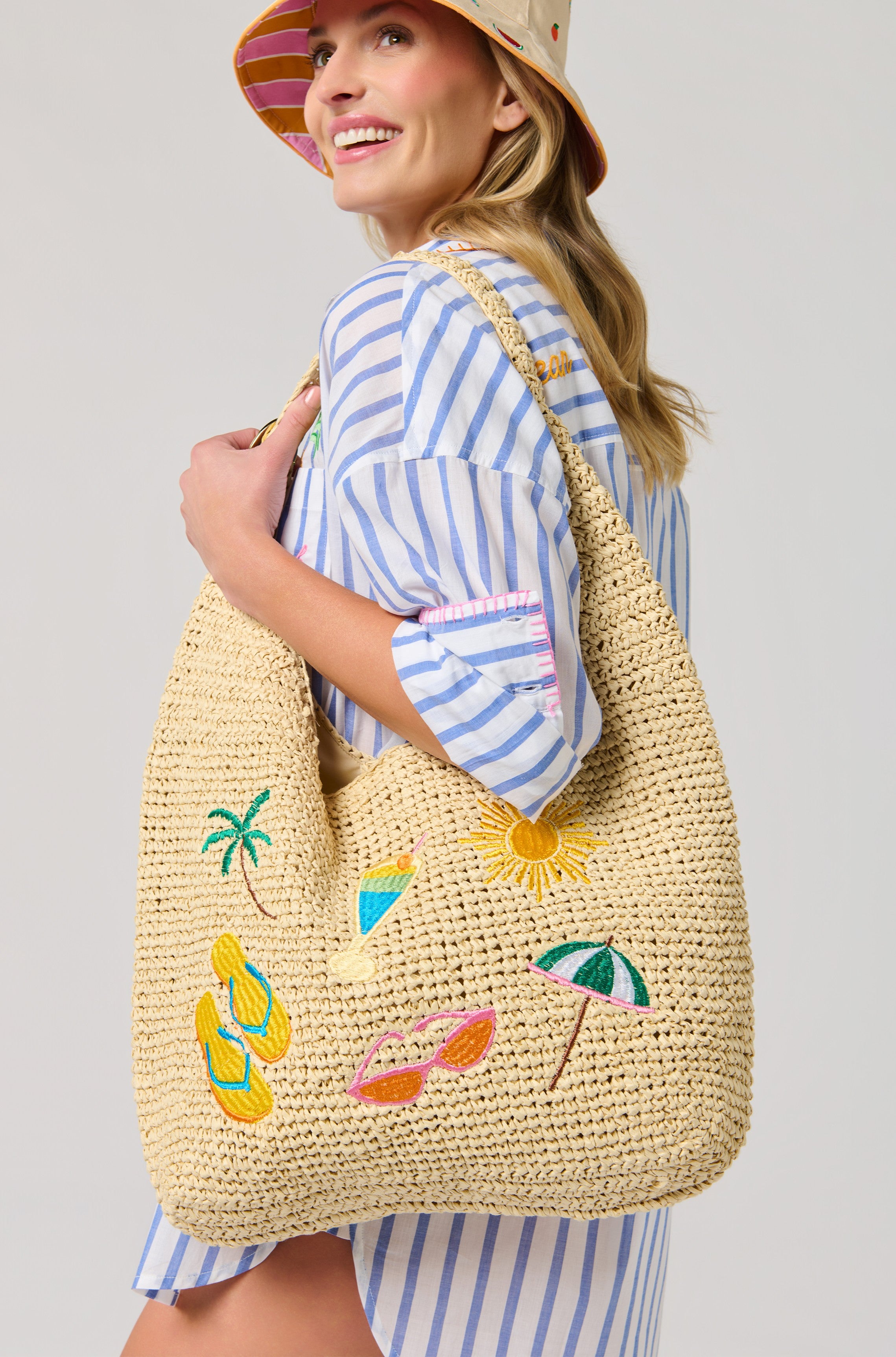 A smiling woman in a blue-and-white striped shirt dress and bucket hat holds the South Moon Under Cabana Slouchy Tote, a colorful beach bag embroidered with sunglasses, flip-flops, palm tree, sun, and umbrella for the ultimate tropical vibe.