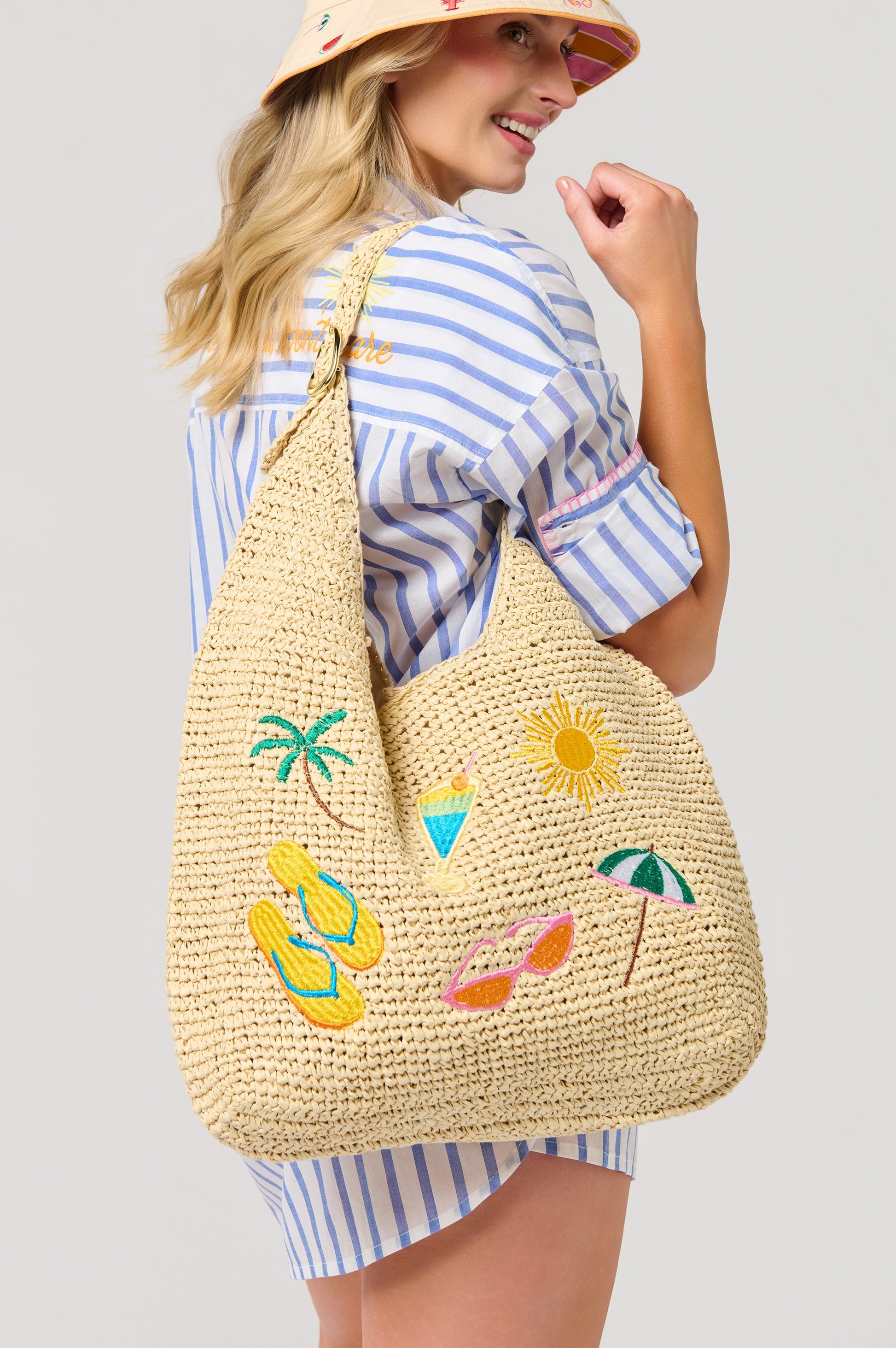 A smiling woman in a striped shirt and sun hat holds the South Moon Under Cabana Slouchy Tote, a vibrant beach bag embroidered with flip-flops, sunglasses, palm tree, cocktail, sun, and umbrella.