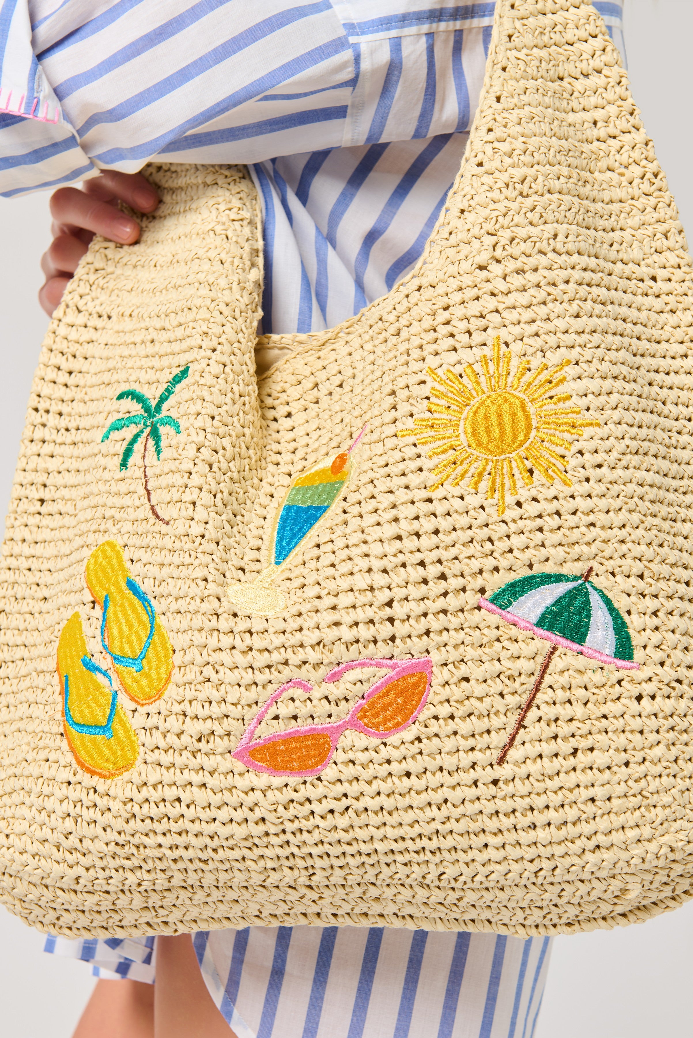 A person in blue-and-white stripes holds the Cabana Slouchy Tote by South Moon Under, a relaxed bag featuring tropical embroidery like a sun, palm tree, drink, flip-flops, sunglasses, and a beach umbrella.