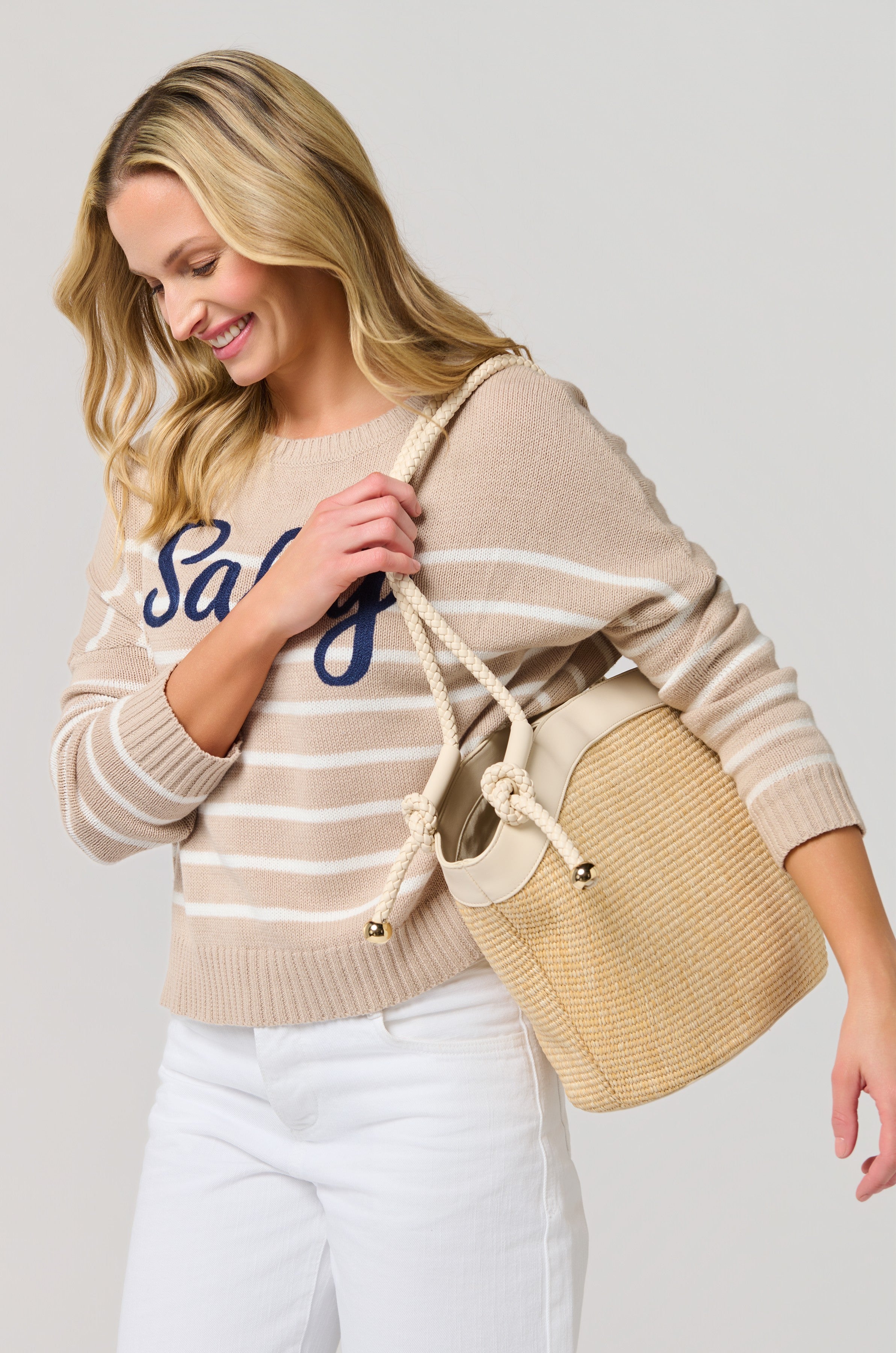 A blonde woman smiles while looking down, wearing a beige and white striped "salty" sweater with white pants and carrying the South Moon Under Gina Tote, featuring faux leather trim.