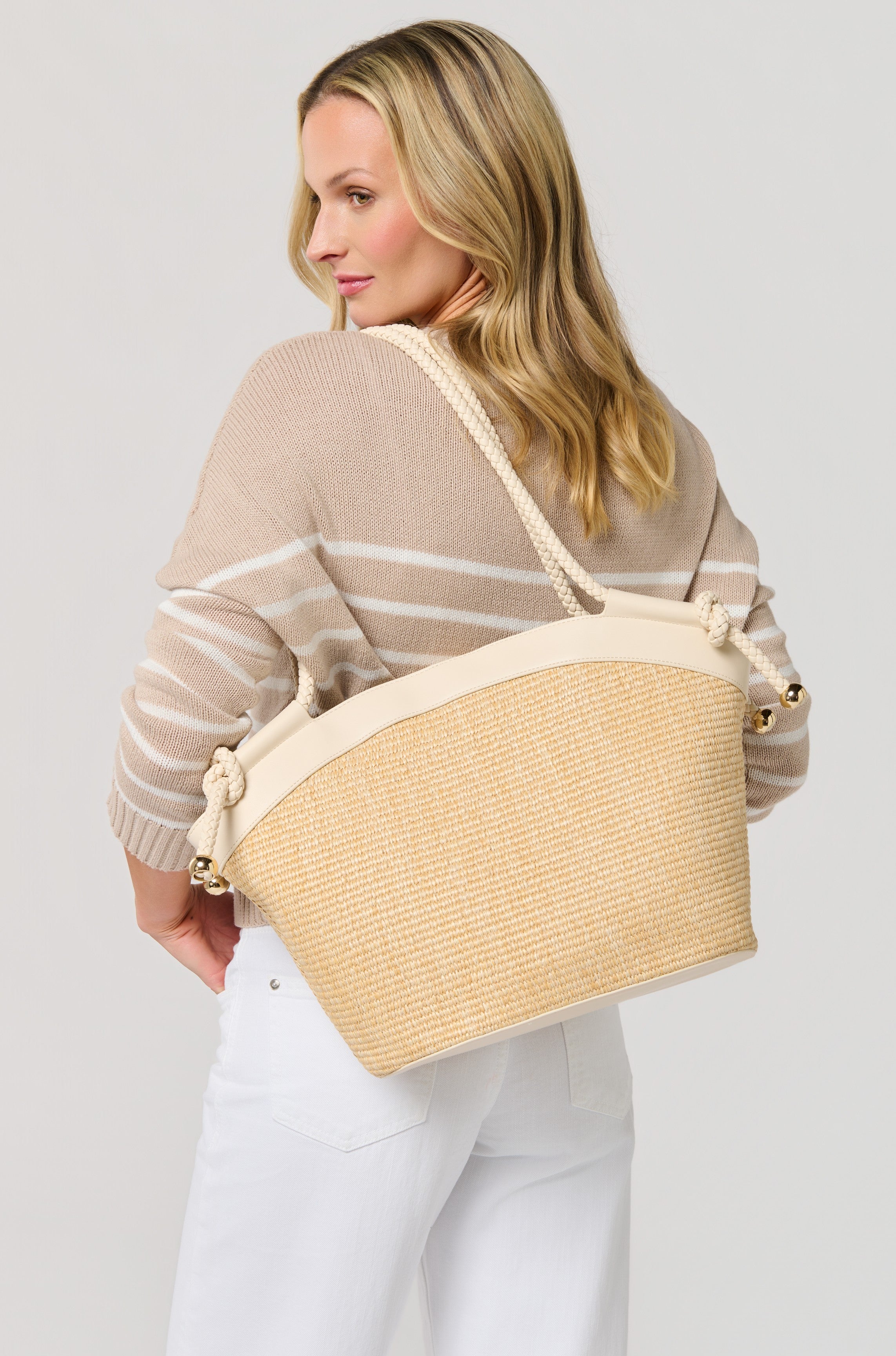 Woman with wavy blonde hair wears a beige striped sweater and white pants, posing with the South Moon Under Gina Tote—a woven bag with faux leather trim and rope handles—over her shoulder, facing slightly away from the camera.