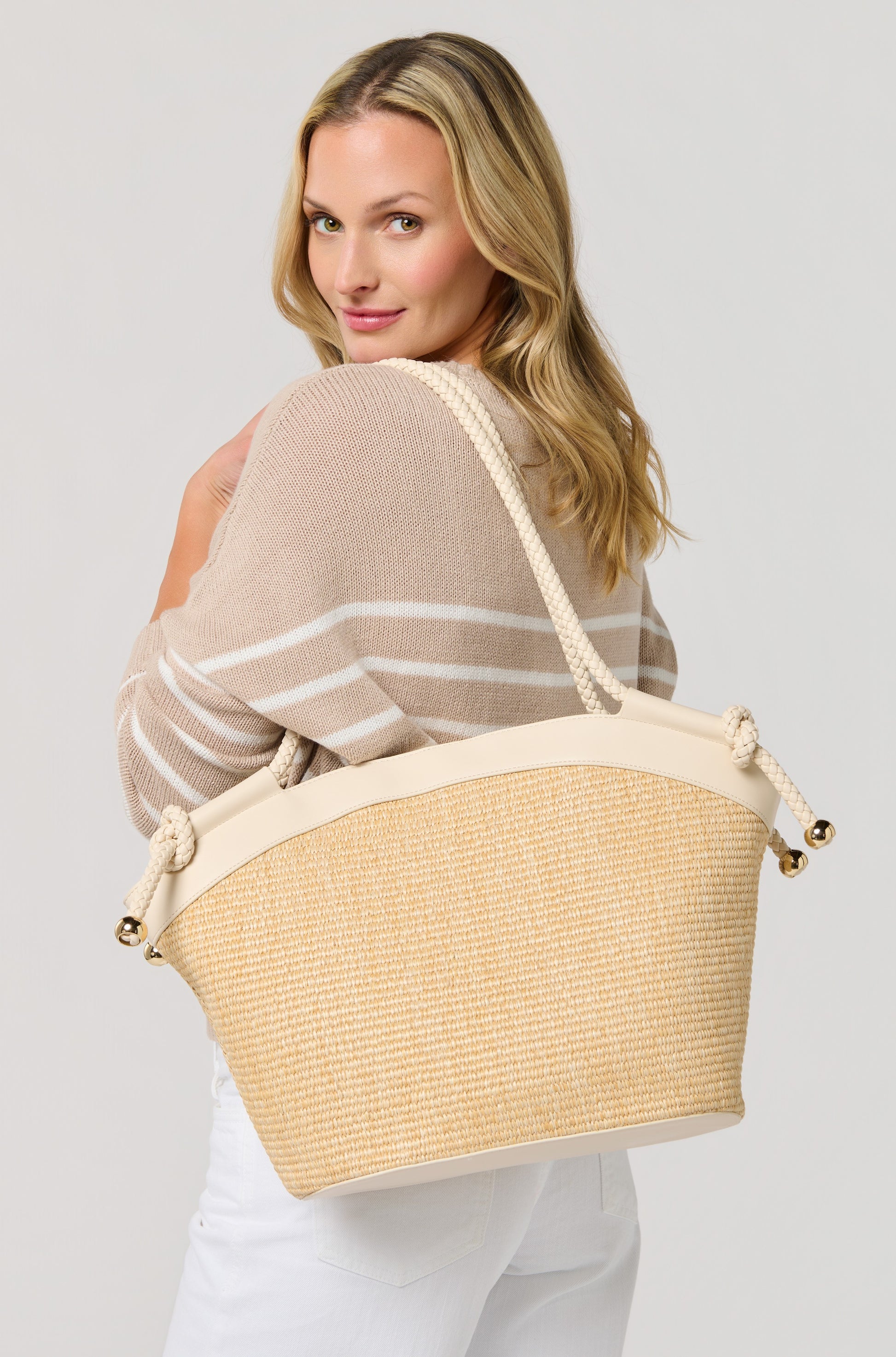A blonde woman smiles over her shoulder, wearing a beige and white striped sweater, white pants, and carrying the South Moon Under Gina Tote in ivory. The background is plain and light-colored.