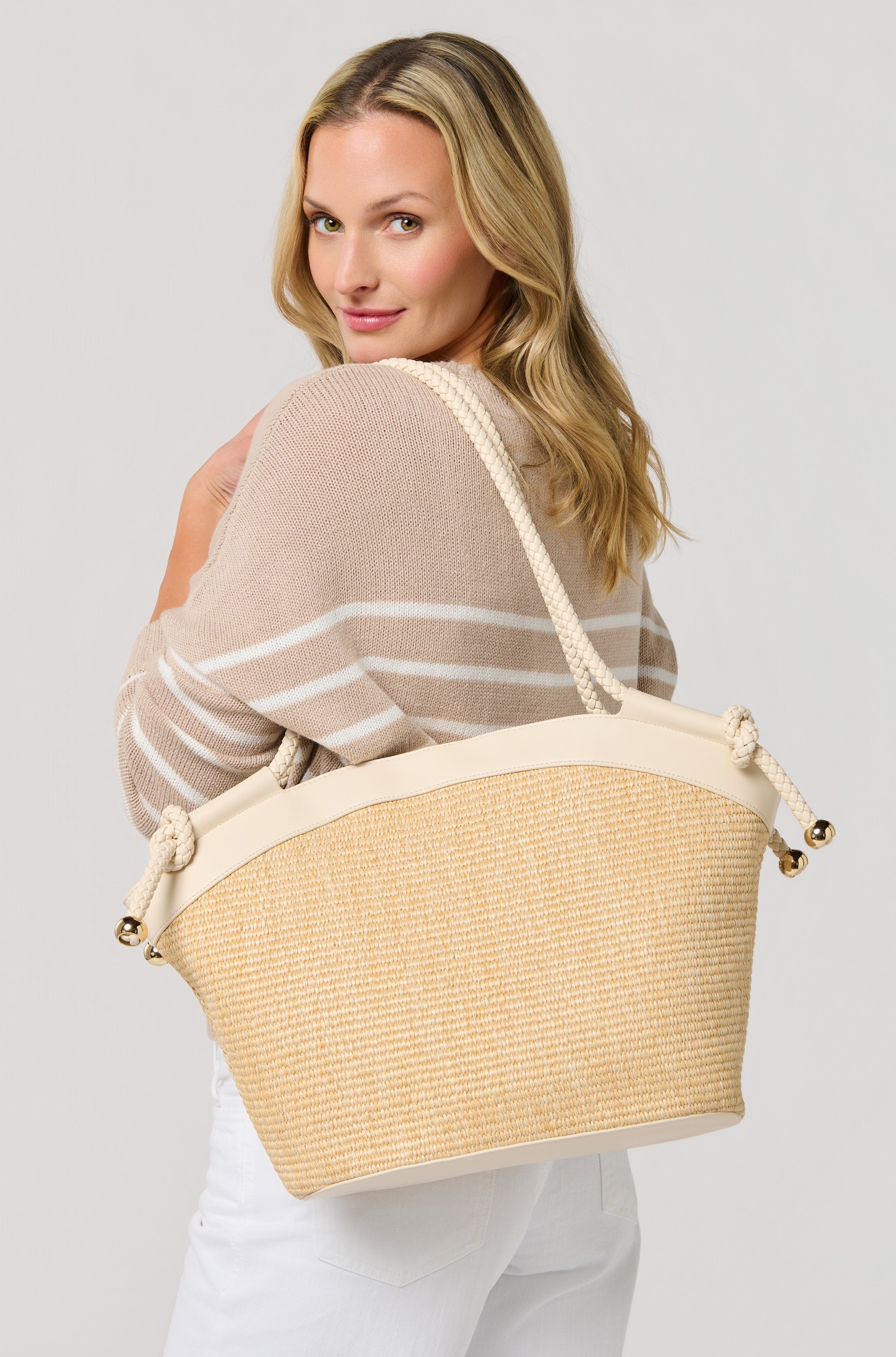 A blonde woman smiles over her shoulder, wearing a beige and white striped sweater, white pants, and carrying the South Moon Under Gina Tote in ivory. The background is plain and light-colored.
