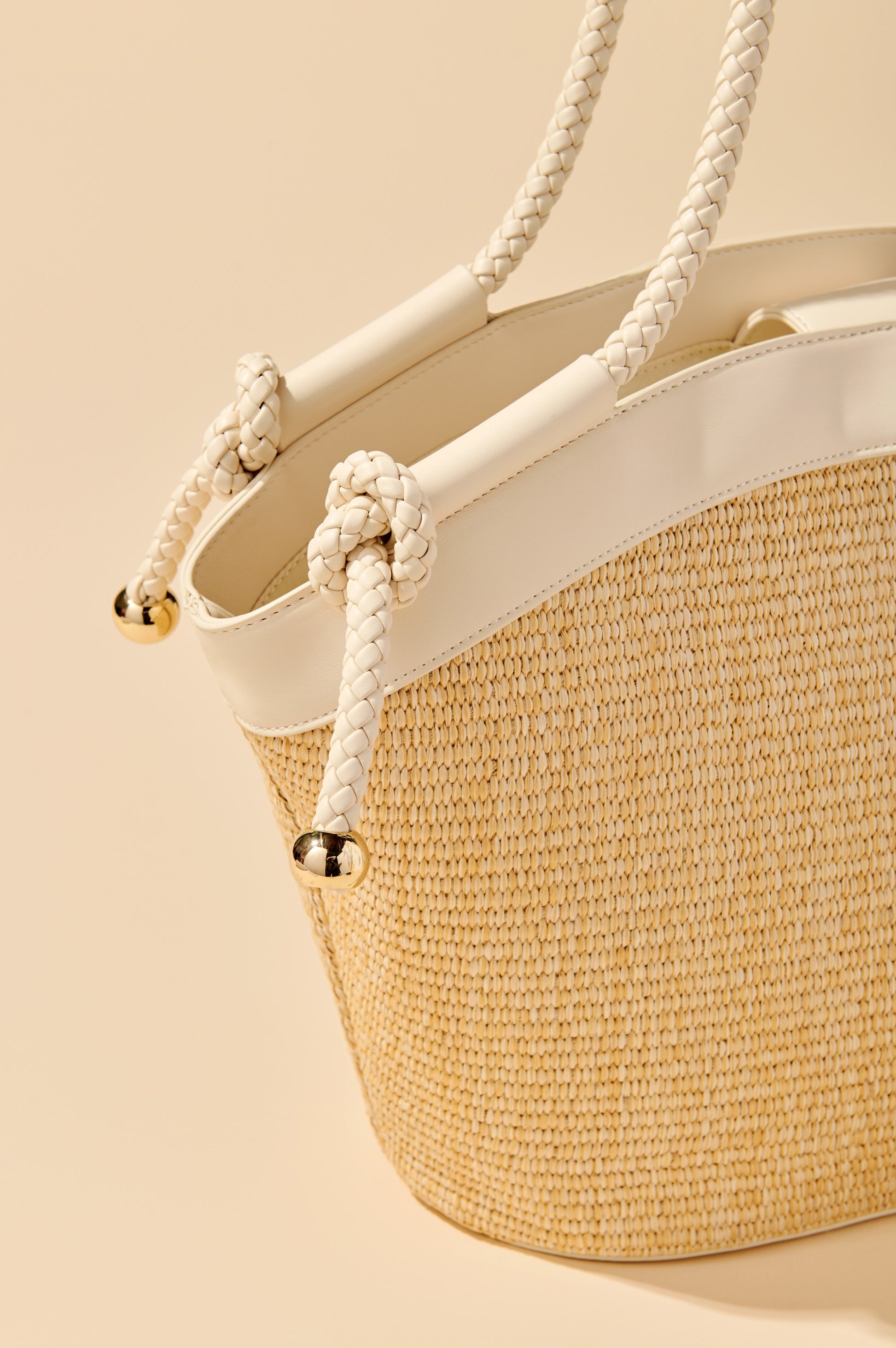A close-up of the Gina Tote by South Moon Under, a woven tote bag with white faux leather trim and braided rope handles featuring gold-tone metal caps, displayed against a beige background.