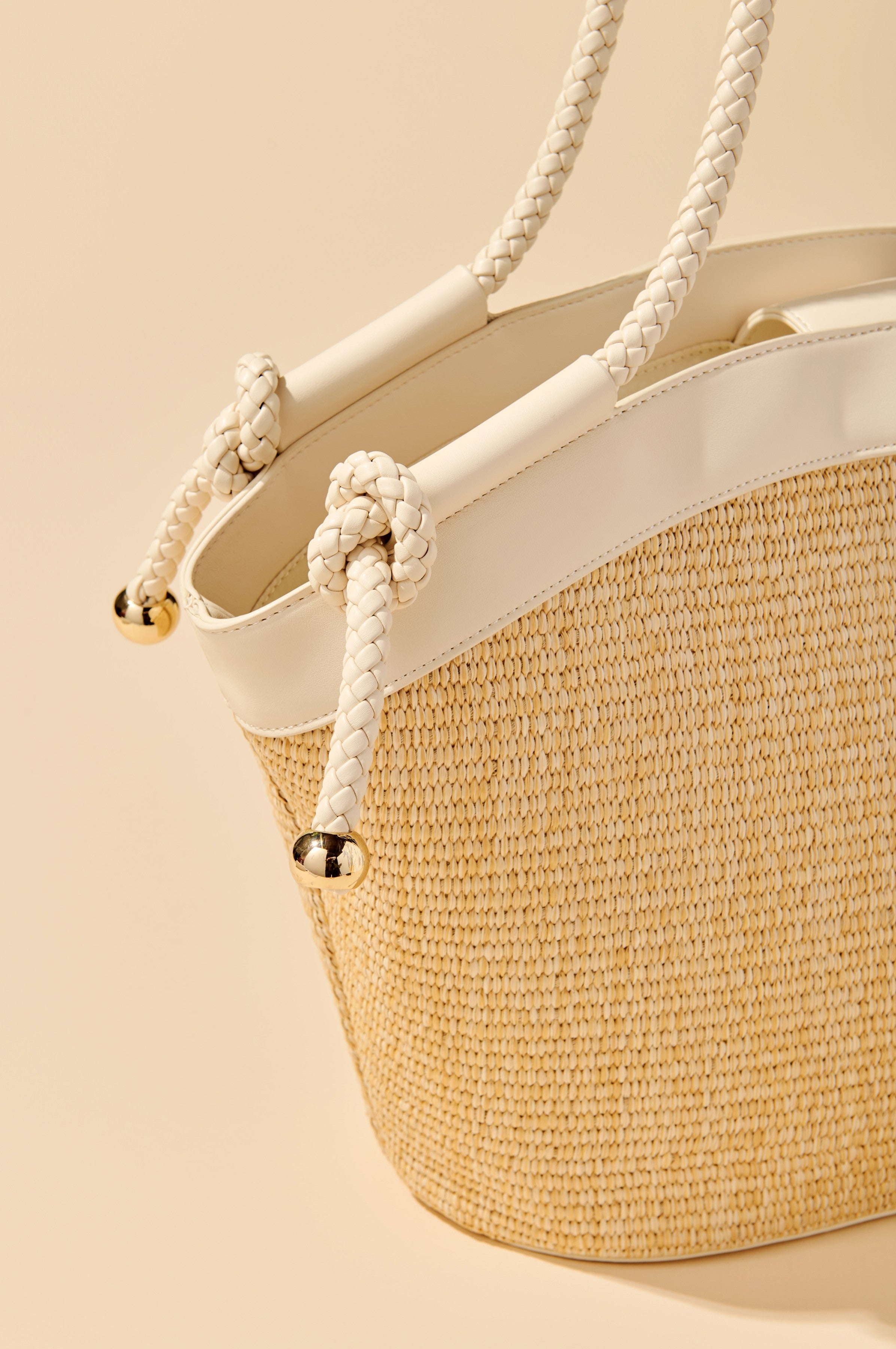 A close-up of the Gina Tote by South Moon Under, a woven tote bag with white faux leather trim and braided rope handles featuring gold-tone metal caps, displayed against a beige background.