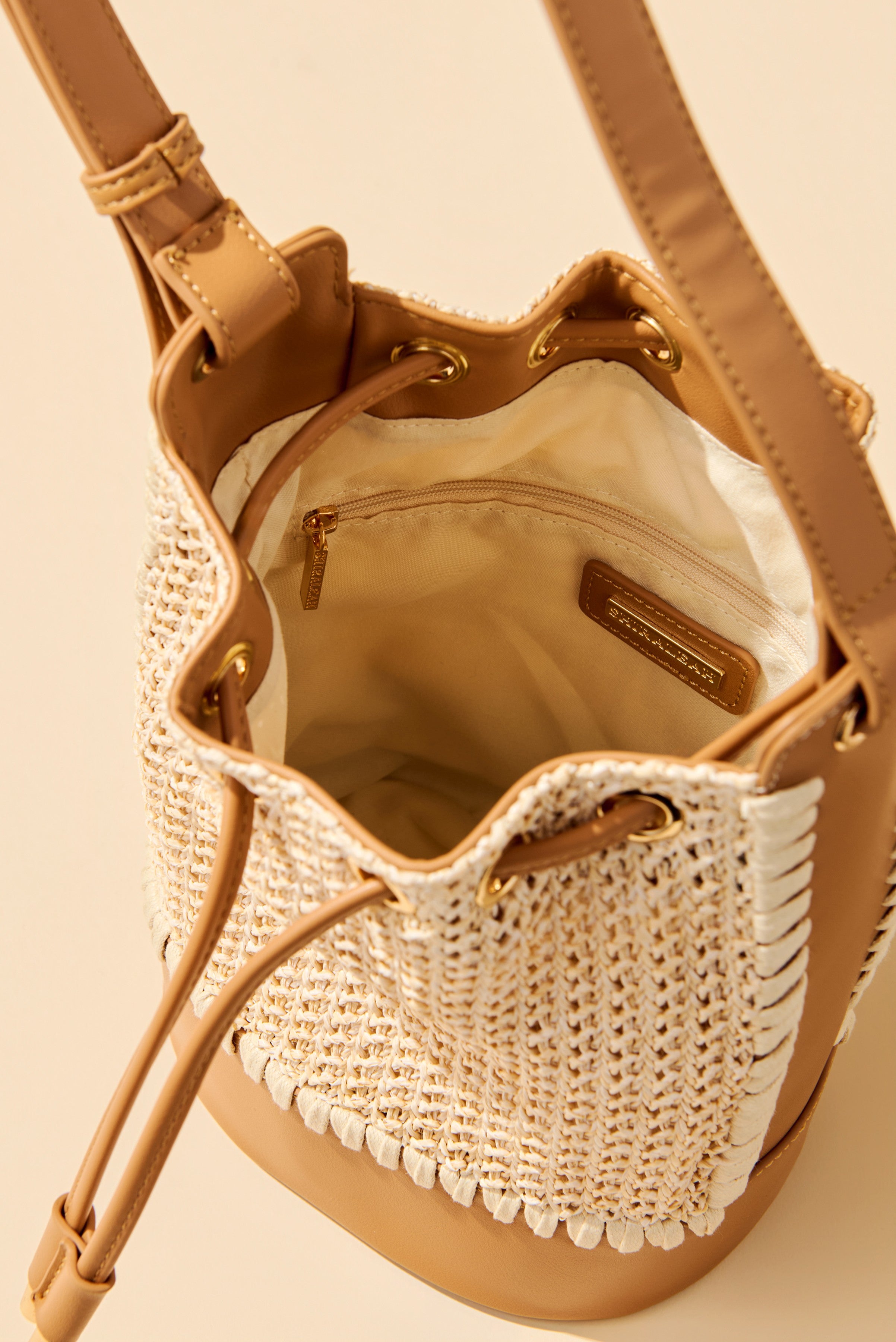 A close-up of the open Mia Bucket Bag by South Moon Under, a tan and white woven summer bag featuring a drawstring closure, gold hardware, and an inner zip pocket—an eye-catching statement accessory against a beige background.