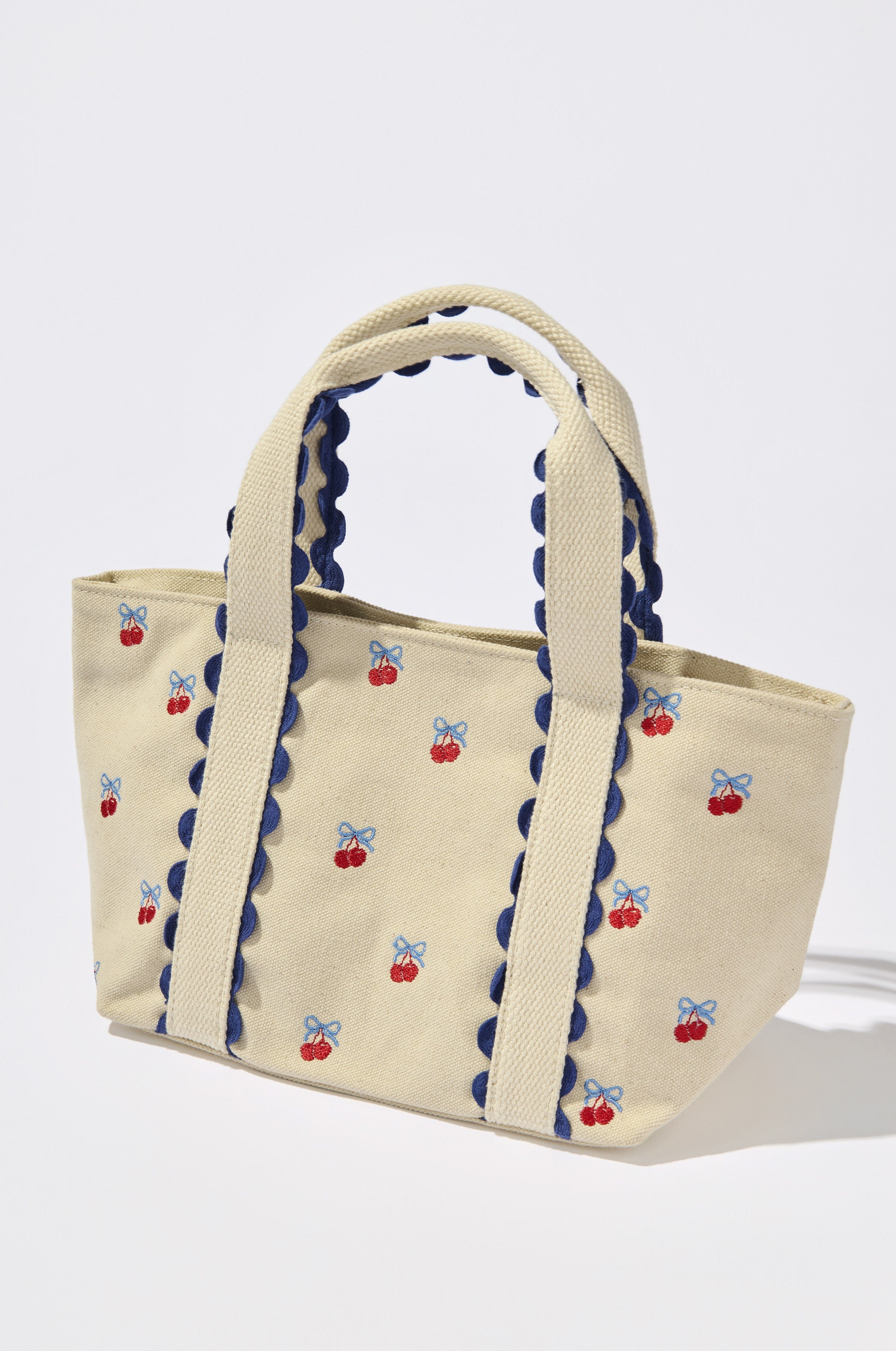 The Cherry Icon Mini Tote by South Moon Under features a Red Multi design with embroidered cherry accents, blue bows, scalloped trim, and sturdy woven handles.