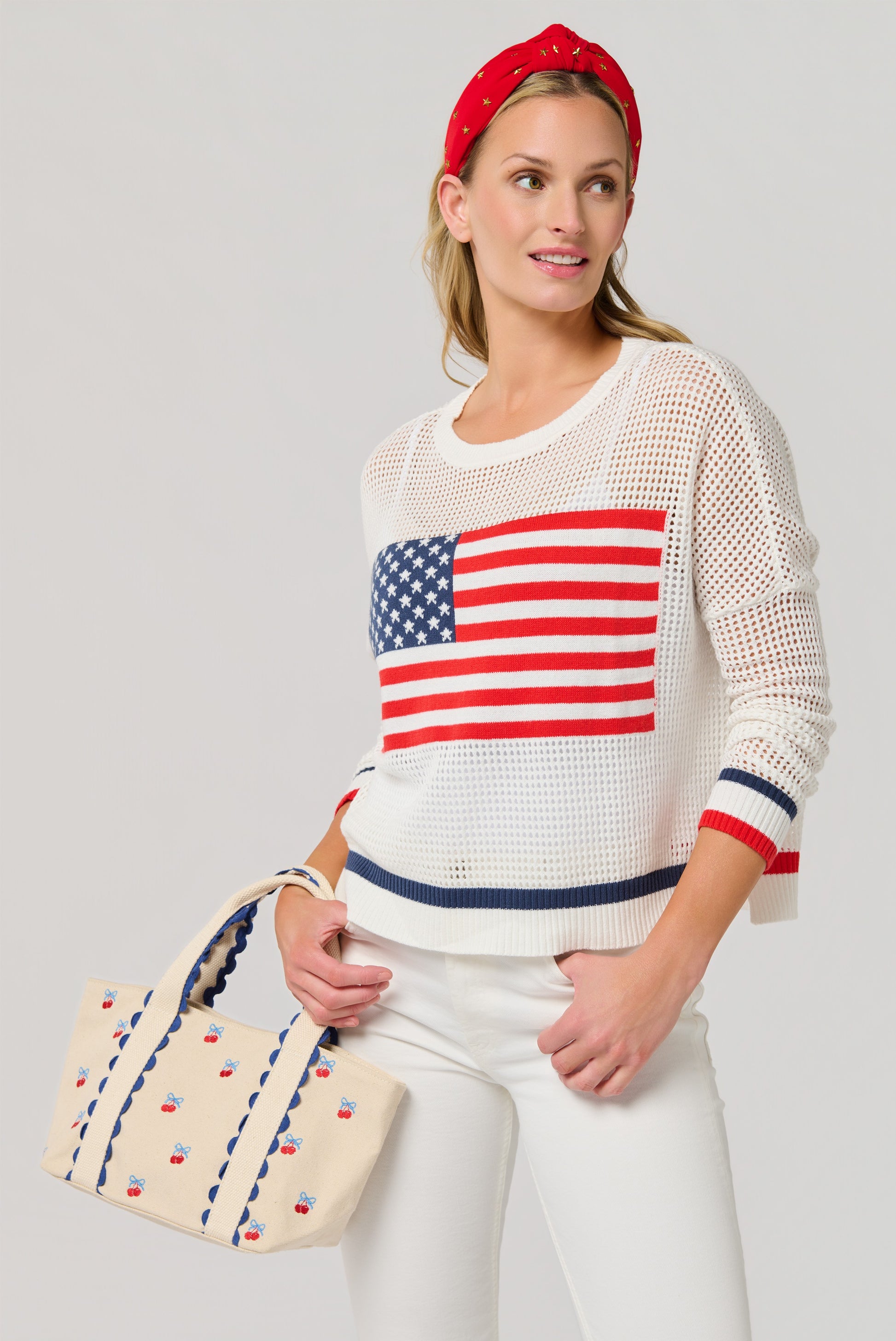 A woman in a white sweater with an American flag design, white pants, and a red headband smiles while holding the Cherry Icon Mini Tote by South Moon Under. She looks to the side against a plain background.