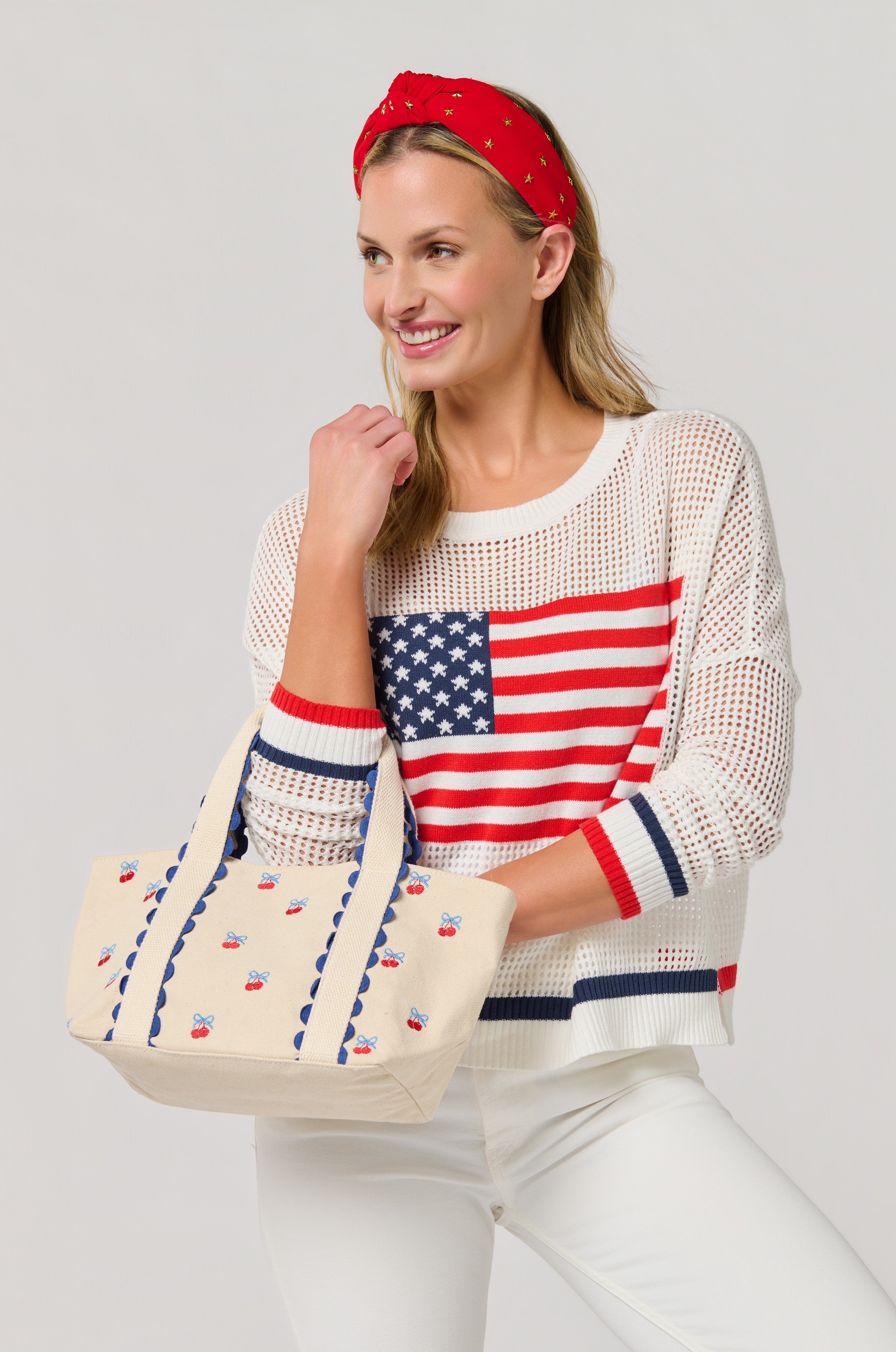 A smiling woman stands against a light background, wearing white pants and a white American flag sweater, holding the South Moon Under Cherry Icon Mini Tote.