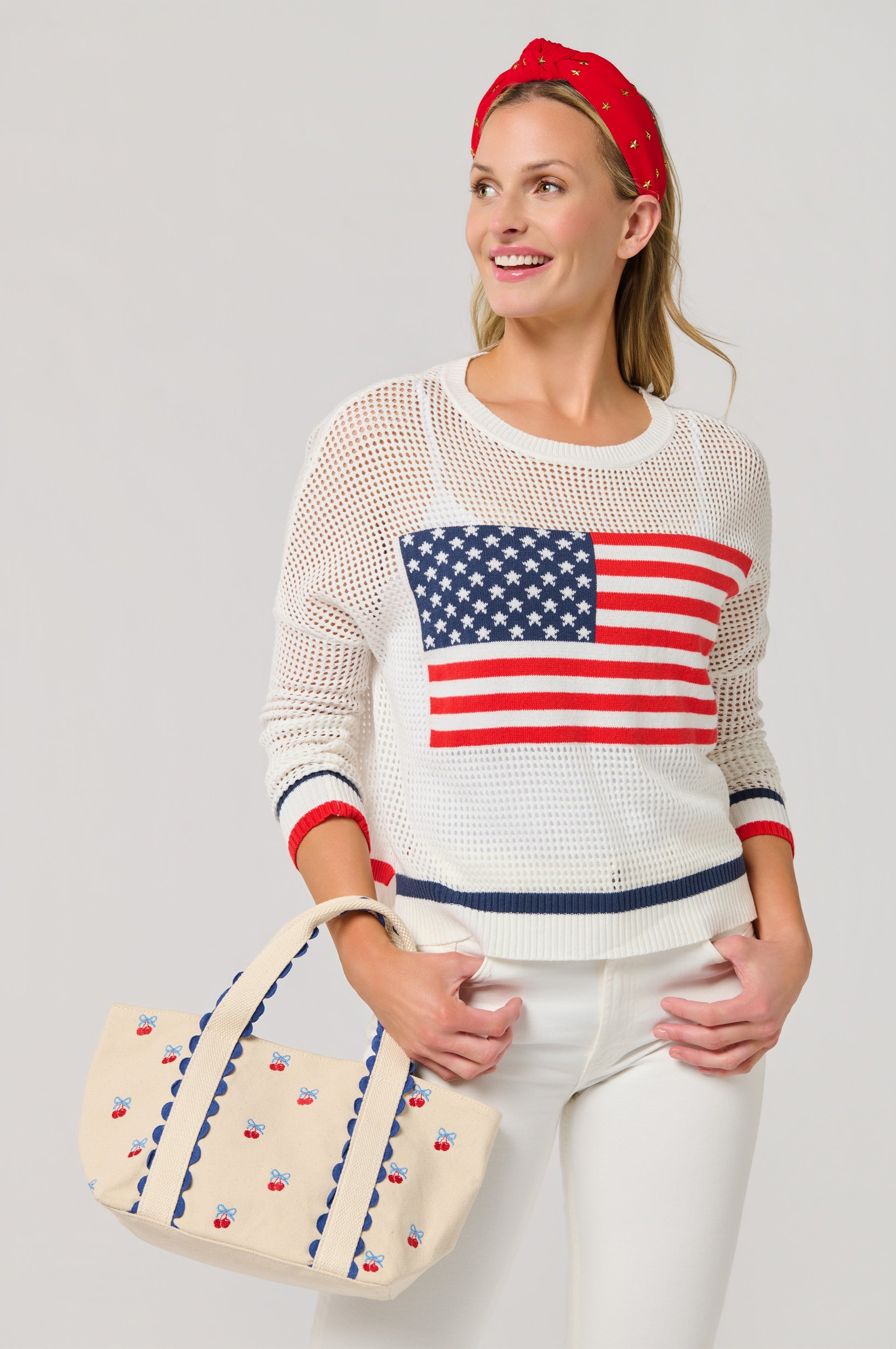A smiling woman wears a white sweater with a large American flag, white pants, and a red headband while holding the South Moon Under Cherry Icon Mini Tote. The tote features embroidered cherries with blue and red accents. Light gray background.