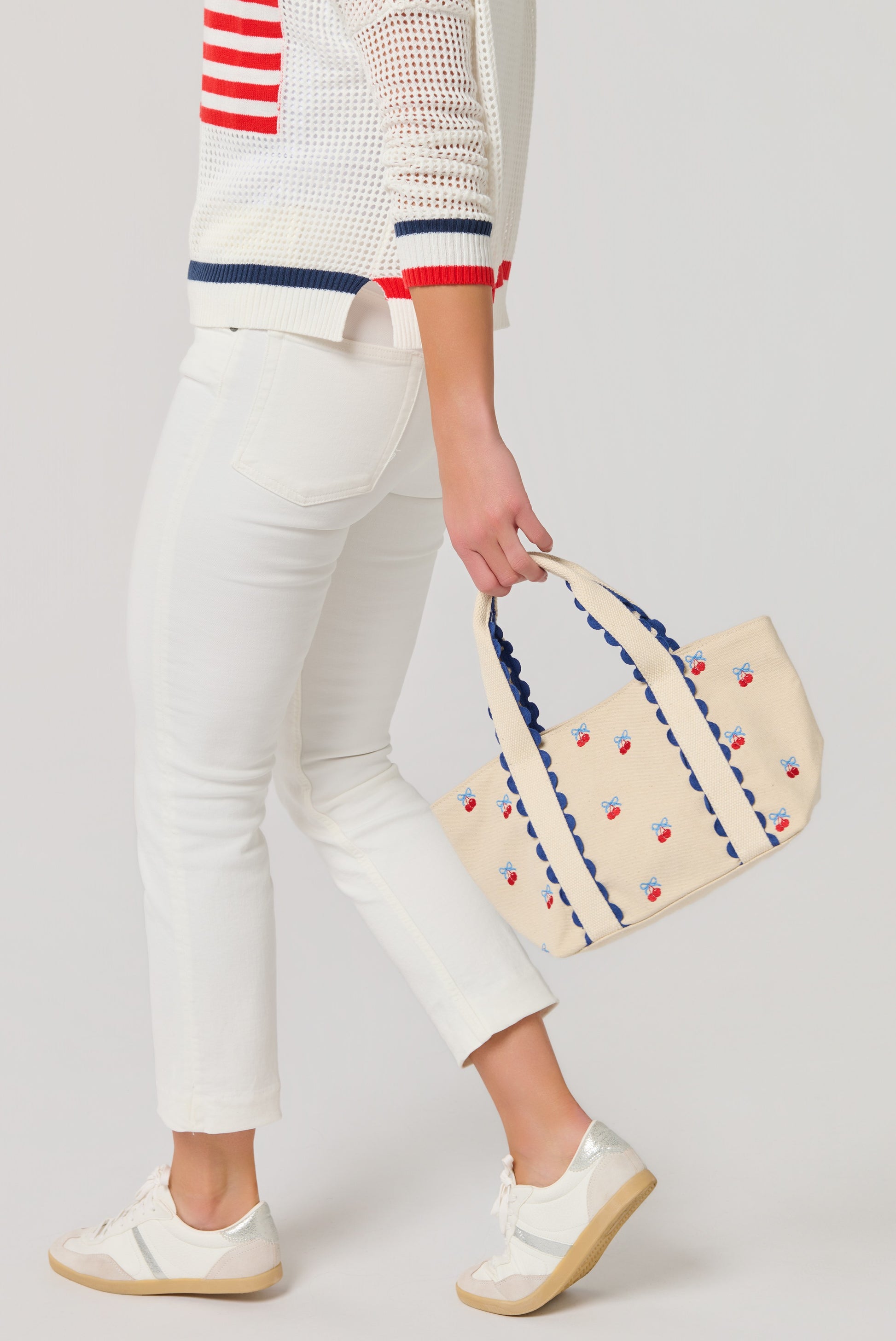 Lower half of person wearing white sweater with red and blue stripes, white pants, and white sneakers, holding the South Moon Under Cherry Icon Mini Tote—a beige canvas bag with small red and blue patterns and scalloped trim.