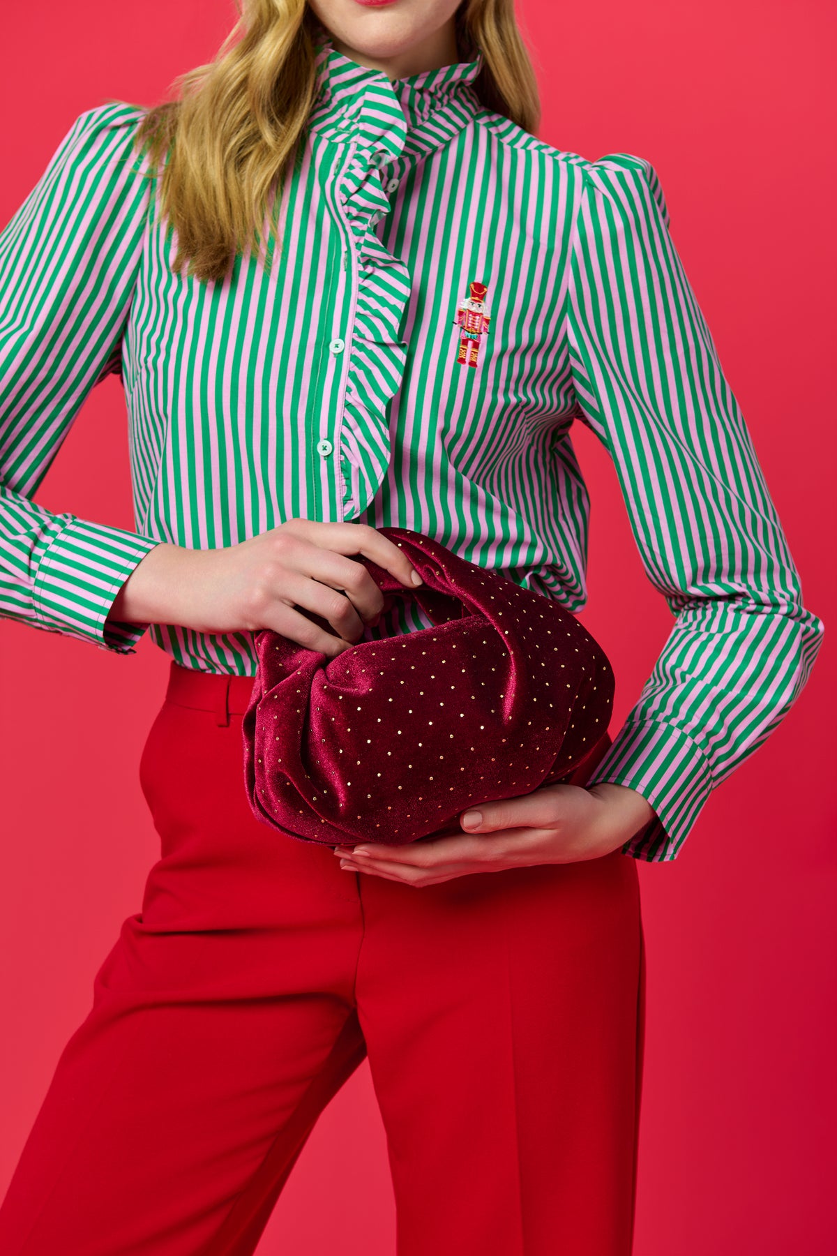 Burgundy velvet top handle bag with studded details, held by a woman against a pink background.