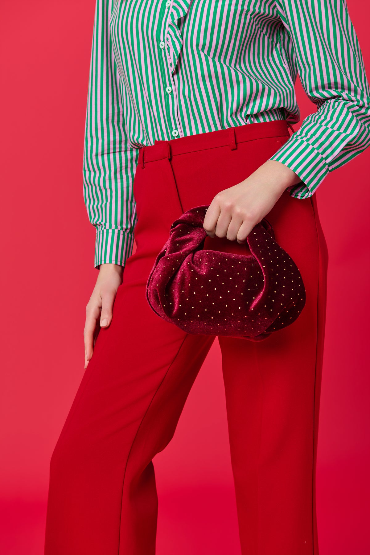 Burgundy velvet top handle bag with gold studded detail, held by model wearing green striped shirt and red pants.