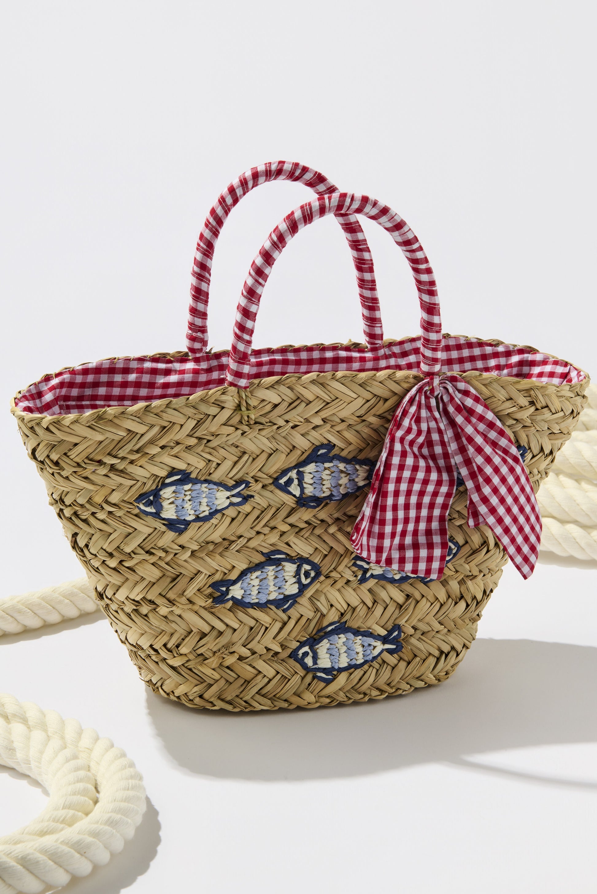 The Marseille Tote by South Moon Under features tan multi woven seagrass with red and white gingham handles, a matching bow, fish embroidery, and gingham lining, shown beside a thick rope for a nautical touch.