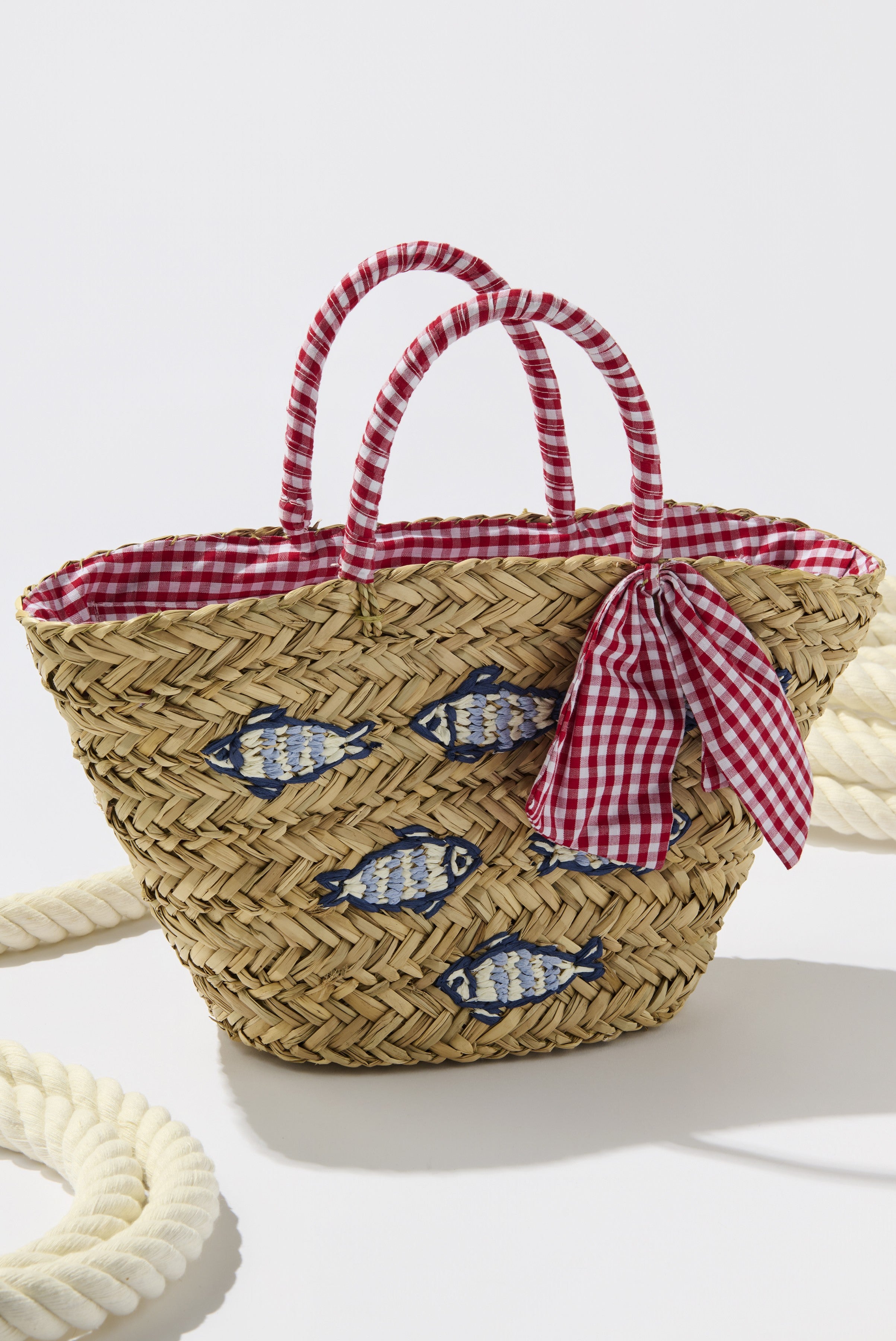 The Marseille Tote by South Moon Under features tan multi woven seagrass with red and white gingham handles, a matching bow, fish embroidery, and gingham lining, shown beside a thick rope for a nautical touch.