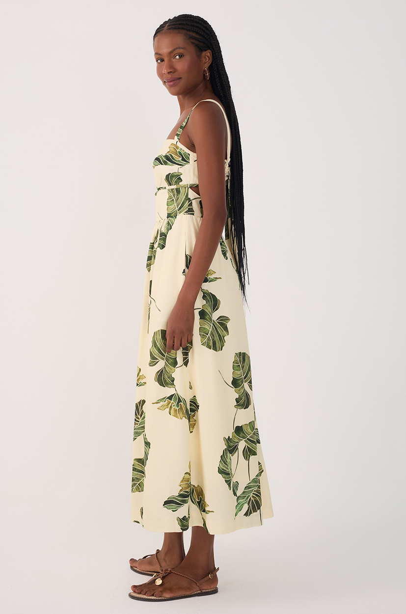 A woman with long braided hair and a subtle smile poses sideways in the Dress To Leaf Print Midi Dress, a sleeveless beige piece featuring green leaves, paired with brown sandals against a plain light background.