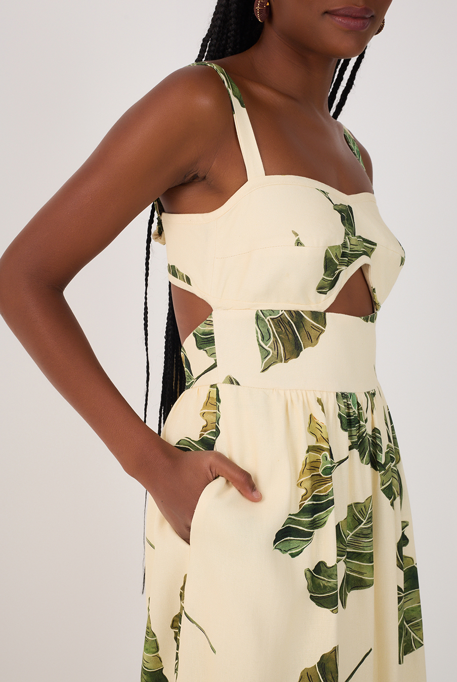 A woman with long braids wears the Dress To Leaf Print Midi Dress, a sleeveless, light yellow cut-out style with green leaf print and pockets. Her hand rests in one pocket; the image is cropped above her knees.