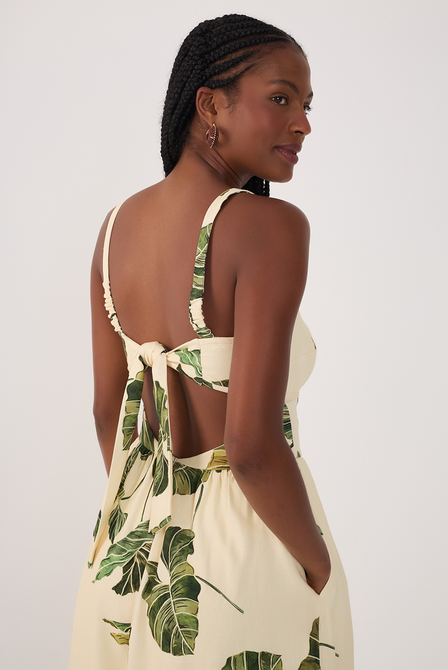 A woman with braided hair wears the Dress To Leaf Print Midi Dress, featuring a green leaf pattern and an open back tied with a bow. She stands sideways with hands in pockets, looking over her shoulder and smiling softly.