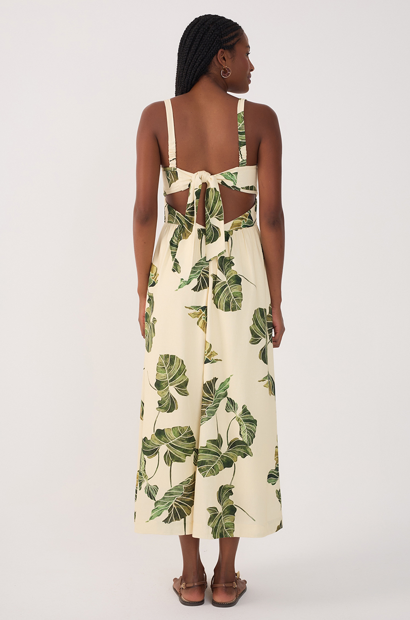 A woman, back to camera, wears the Dress To Leaf Print Midi Dress—a sleeveless, cream midi with green leaf prints, wide straps, and an open back with bow tie—paired with sandals.