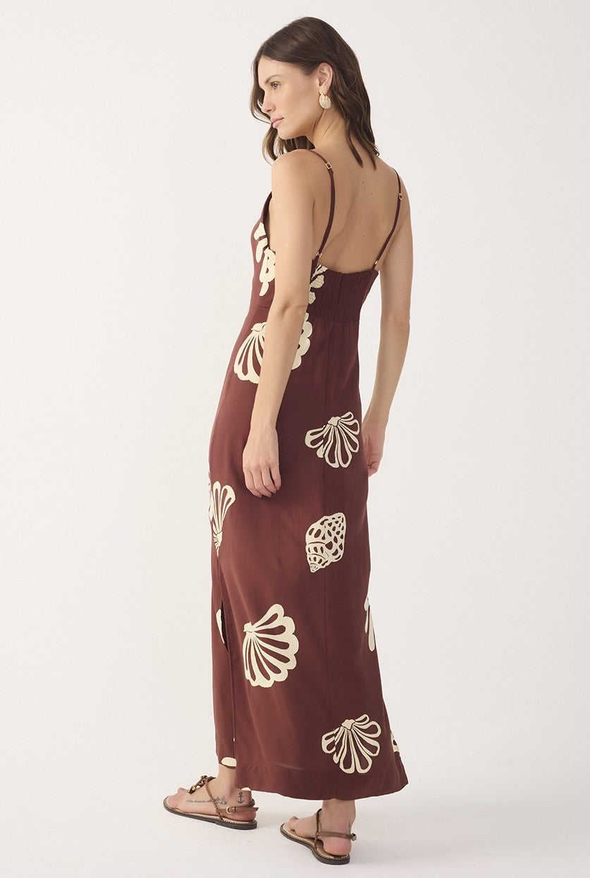 A woman stands barefoot, turned slightly to the side, wearing the Dress To Brown Maritime Cut-out Smocked Maxi Dress featuring adjustable spaghetti straps and white shell and sea motifs, against a plain white background.