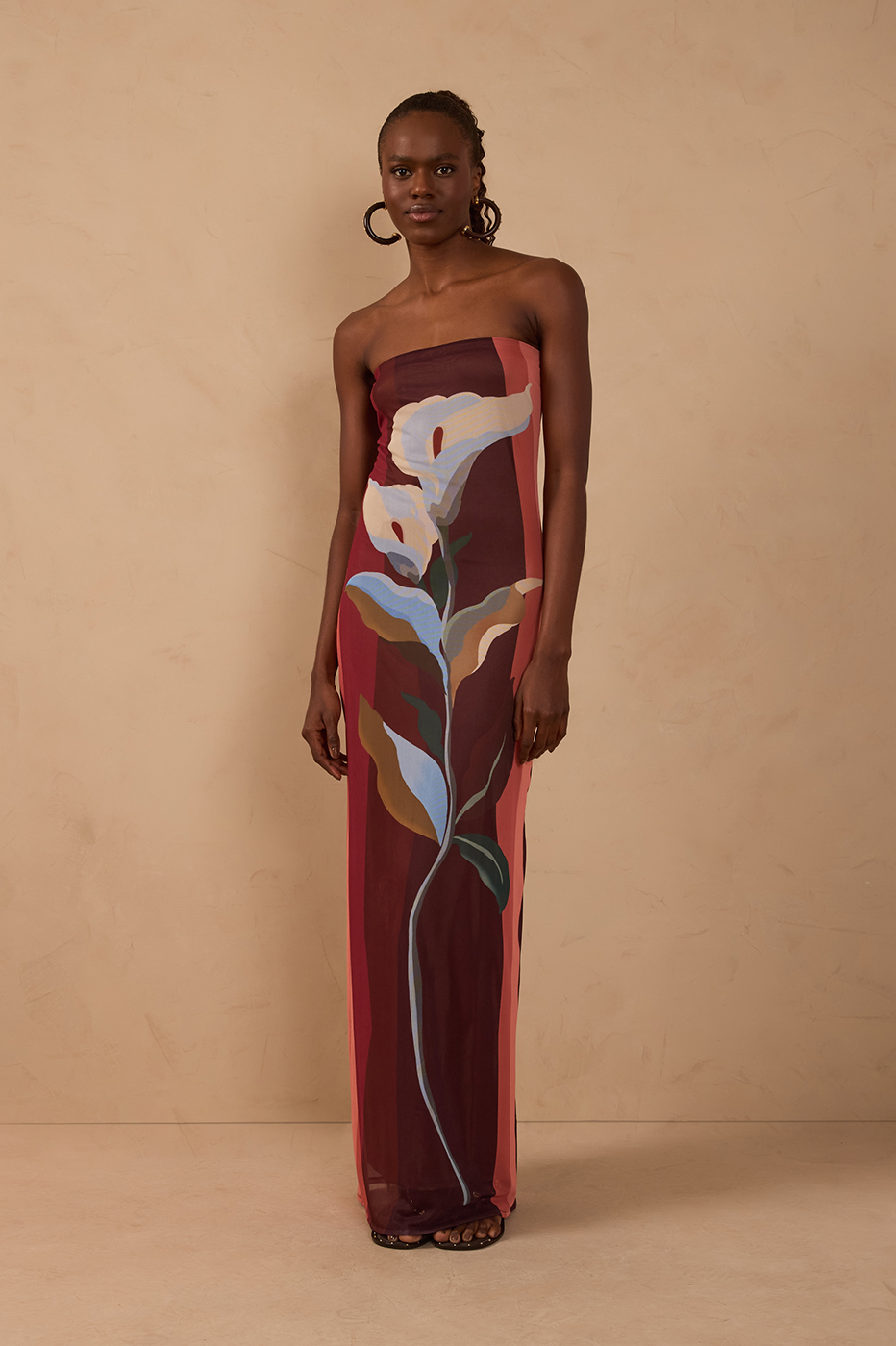 Against a beige background, a woman wears the Dress To Multicolor B Risa Strapless Tulle Maxi in MULTICOLOR BRISA, a floor-length dress featuring a vibrant botanical print and sheer overlay for airy elegance, styled with hoop earrings—an ideal choice for resort wear.