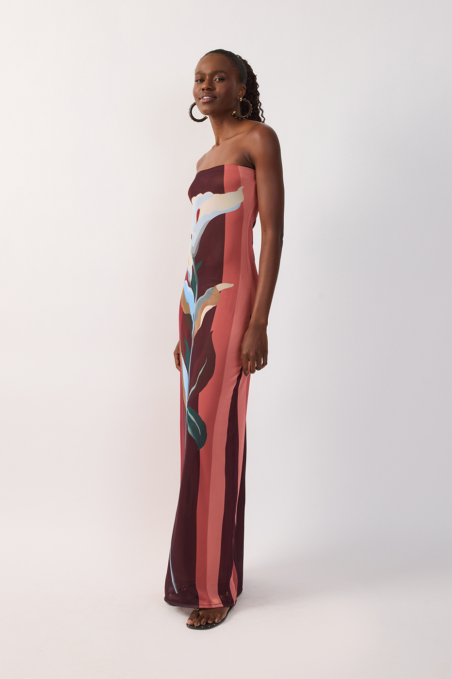 A woman wearing the Dress To Multicolor B Risa Strapless Tulle Maxi, a floor-length dress featuring a bold abstract floral print in burgundy, pink, and blue tones with a delicate sheer overlay, stands against a plain white background.