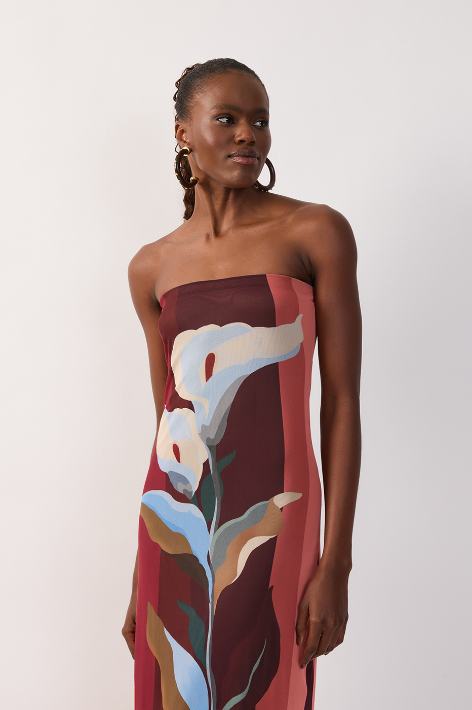 Woman standing against a plain background, wearing the Dress To Multicolor B Risa Strapless Tulle Maxi, featuring a vibrant botanical print in white, blue, and green tones. She accessorizes with hoop earrings and wears her hair styled back, radiating effortless resort wear elegance.