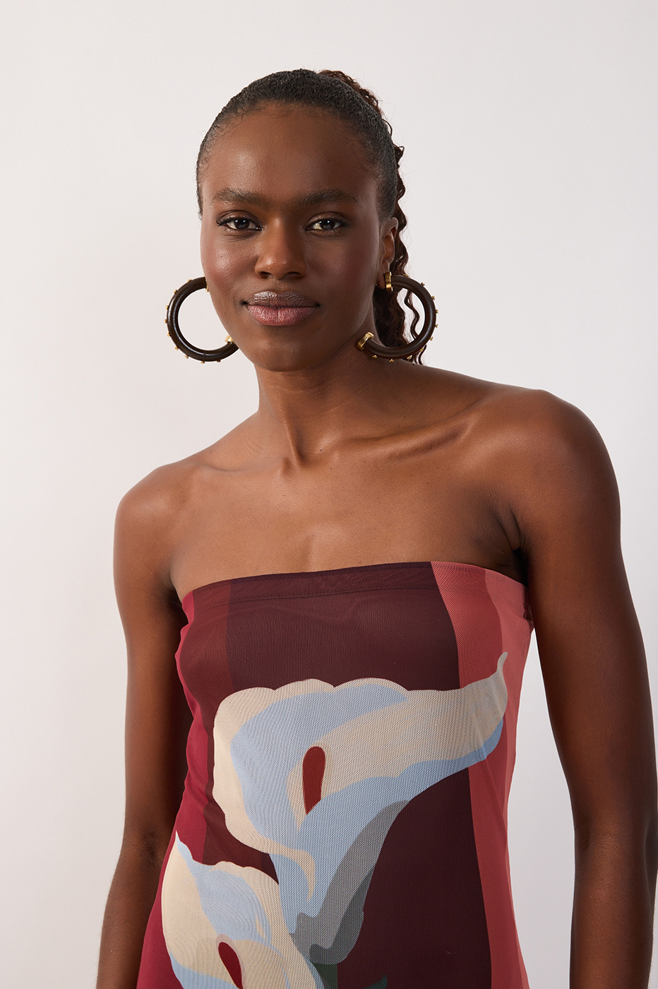 A woman with hoop earrings and braided hair wears the Dress To Multicolor B Risa Strapless Tulle Maxi, featuring a large floral print, as she stands against a plain white background.