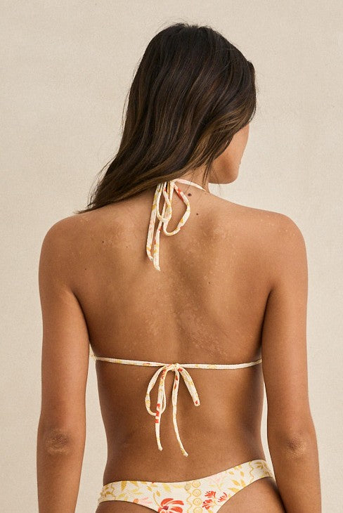A woman with long brown hair stands facing away against a beige background, wearing the Rhythm Wildflower Slide Tri Top—a fully lined floral bikini with adjustable string ties at the neck and back.