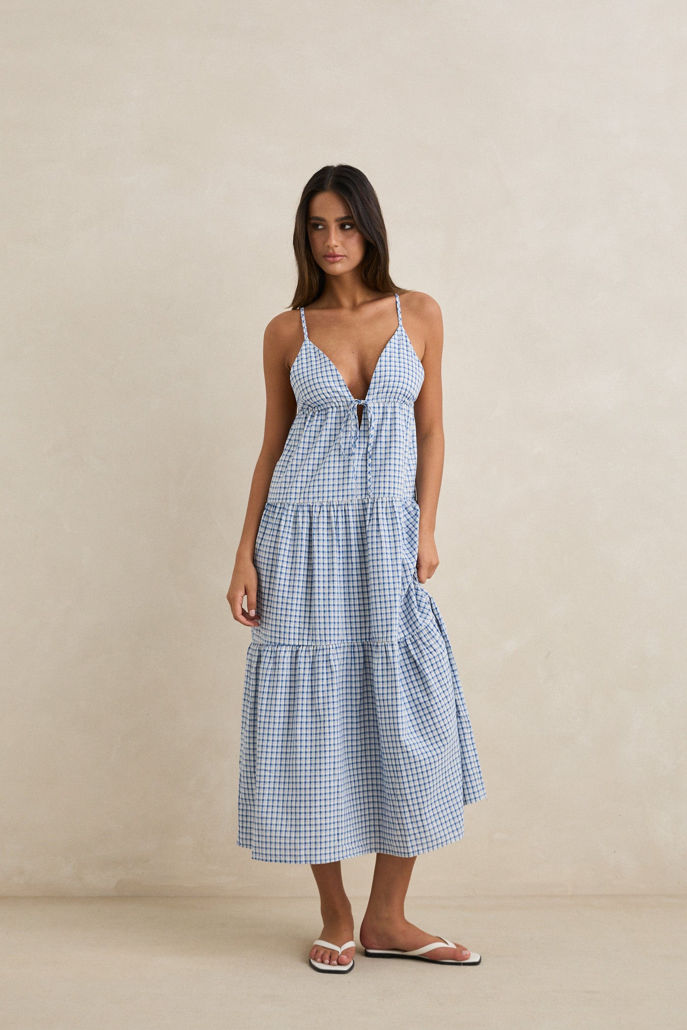 A woman in the Rhythm Jaci Maxi Dress—a blue and white gingham 100% cotton sleeveless maxi with spaghetti straps—stands against a plain beige background, looking slightly to the side and wearing white sandals.