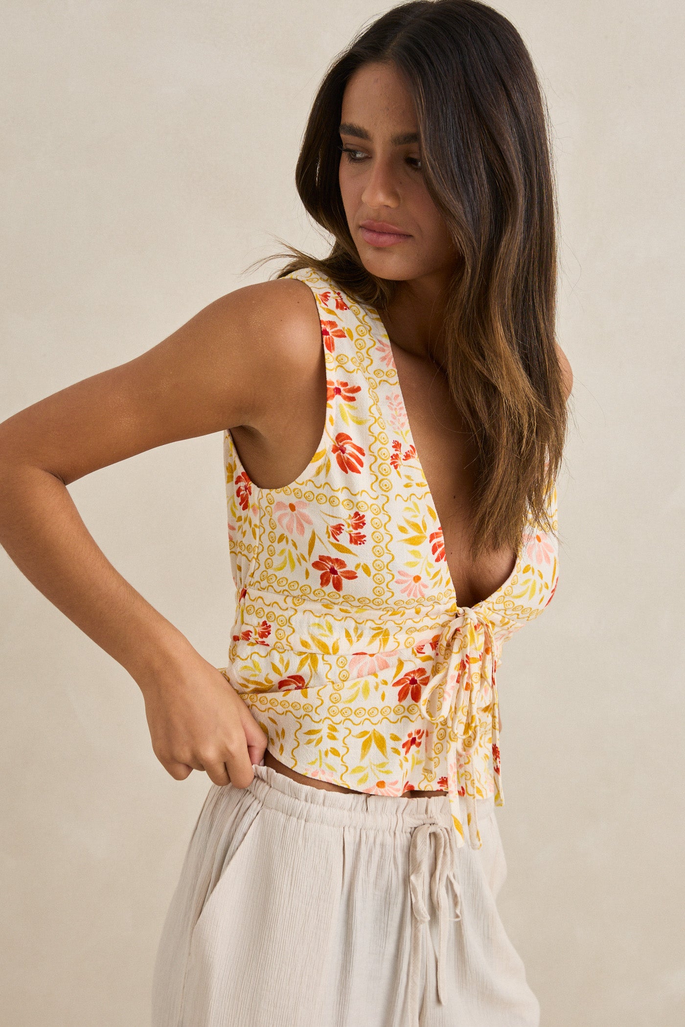A woman with long brown hair models the Rhythm Wildflower Tie Front Top, a sleeveless v-neck style with a yellow and red floral print and adjustable tie front, paired with cream drawstring pants against a neutral background.