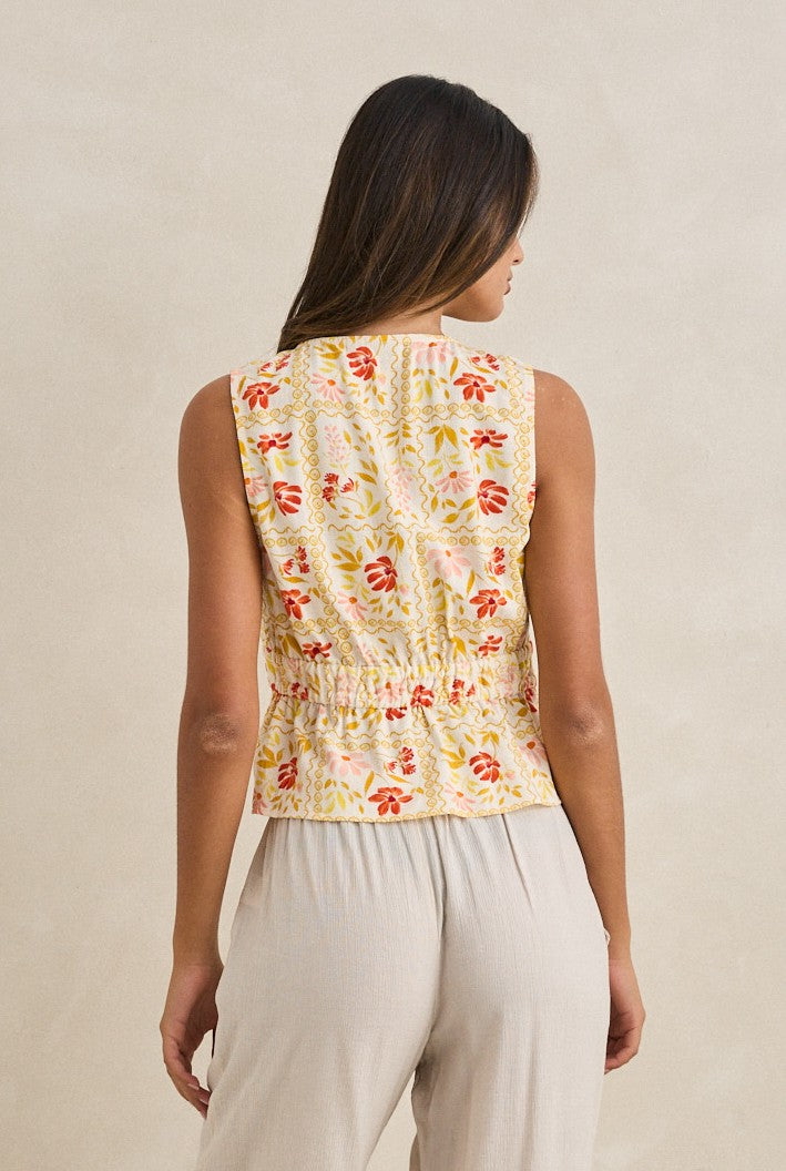 A woman with long, dark hair faces away, wearing the Rhythm Wildflower Tie Front Top—a sleeveless yellow floral piece with red and green details—and light beige pants, set against a plain cream background.