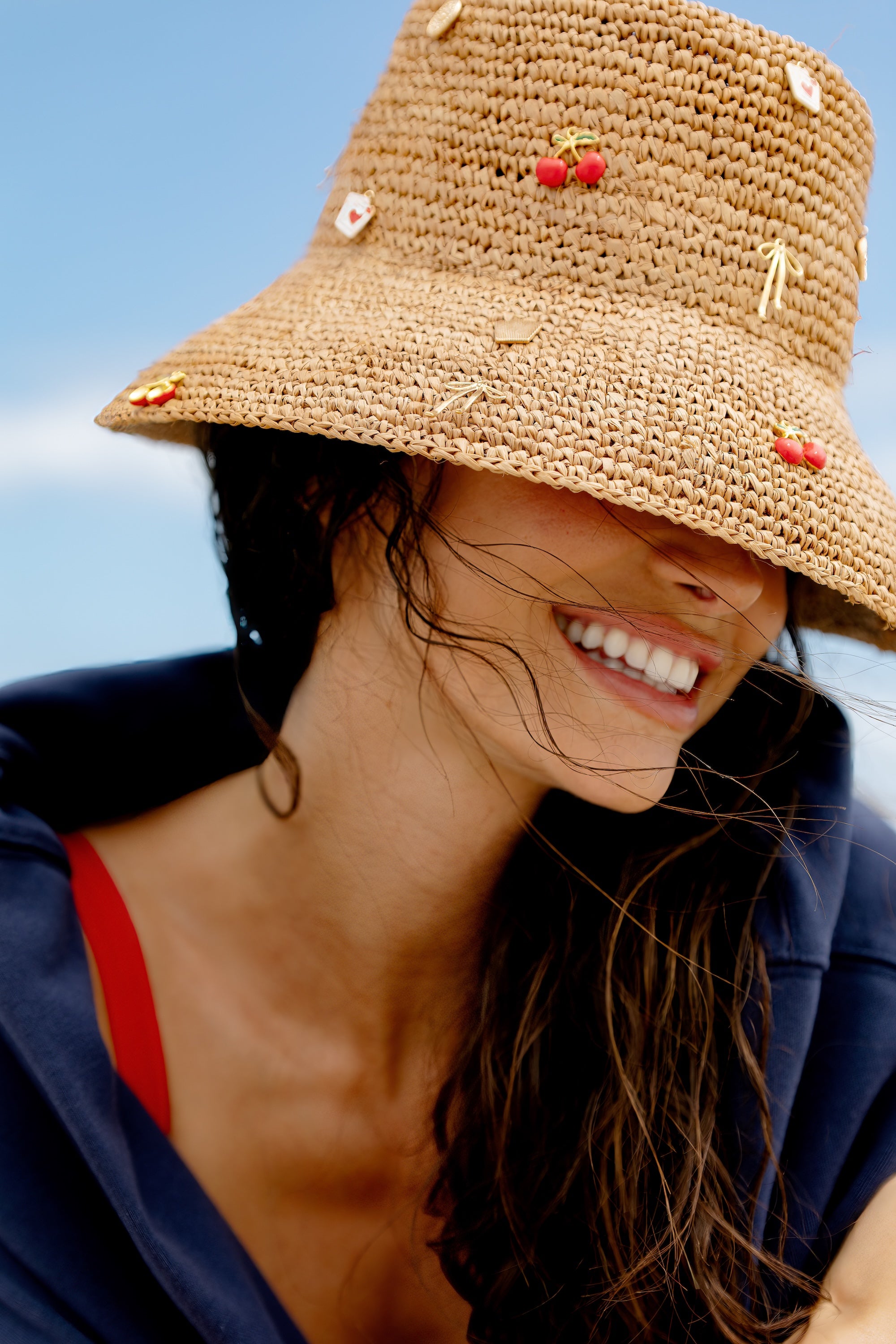 A woman smiles brightly, wearing the San Diego Hat Co. Charm Bucket Hat in tan, featuring UPF 50+ sun protection and cherry accents. Her windblown dark hair and navy top with a red strap stand out against the blue sky backdrop.