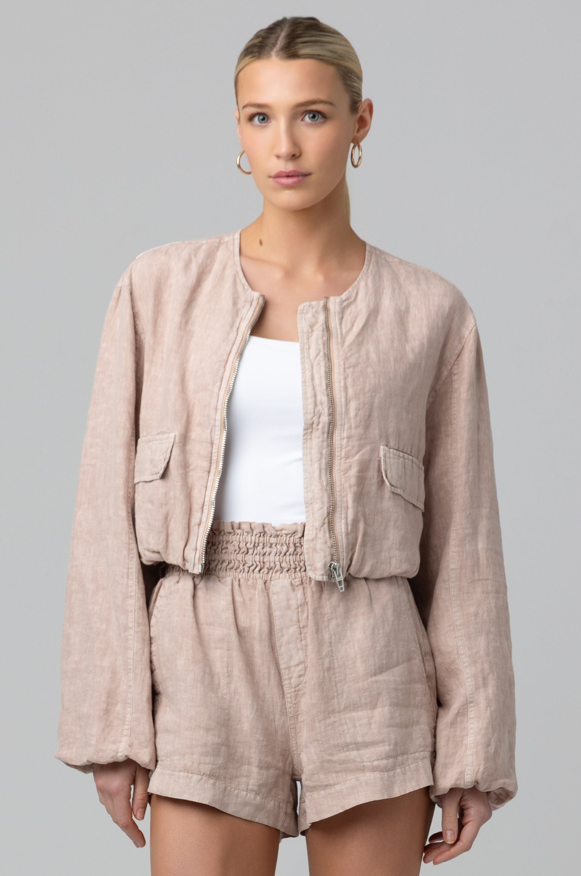 A woman wears the Blank Read My Lips Jacket in light pink, a 100% linen collarless bomber, styled with matching high-waisted shorts and a white top. Her blonde hair is tied back as she stands against a plain gray background.