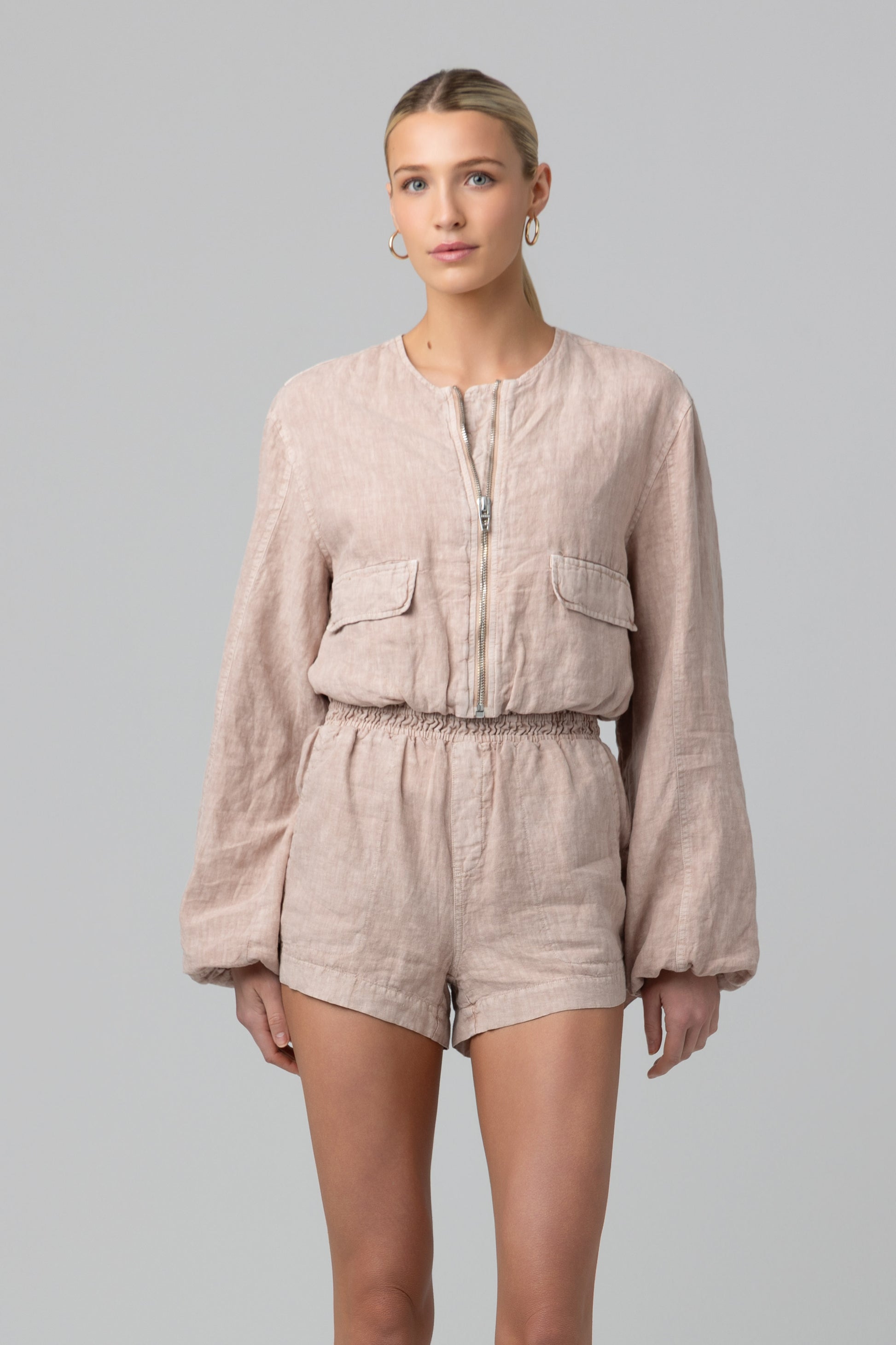 A woman with her hair tied back and hoop earrings models the Read My Lips Jacket by Blank—a collarless, 100% linen, long-sleeve zip-up romper featuring two front pockets and an elastic waistband, set against a plain background.