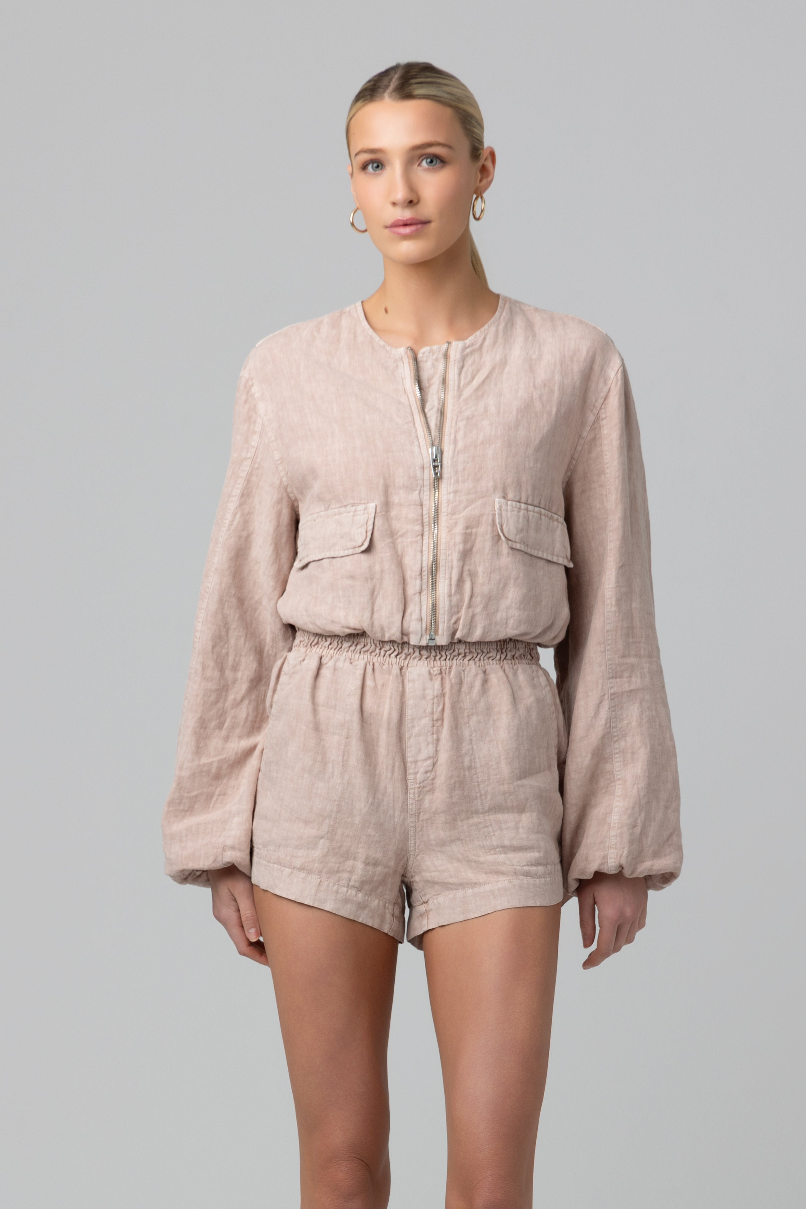 A woman with her hair tied back and hoop earrings models the Read My Lips Jacket by Blank—a collarless, 100% linen, long-sleeve zip-up romper featuring two front pockets and an elastic waistband, set against a plain background.