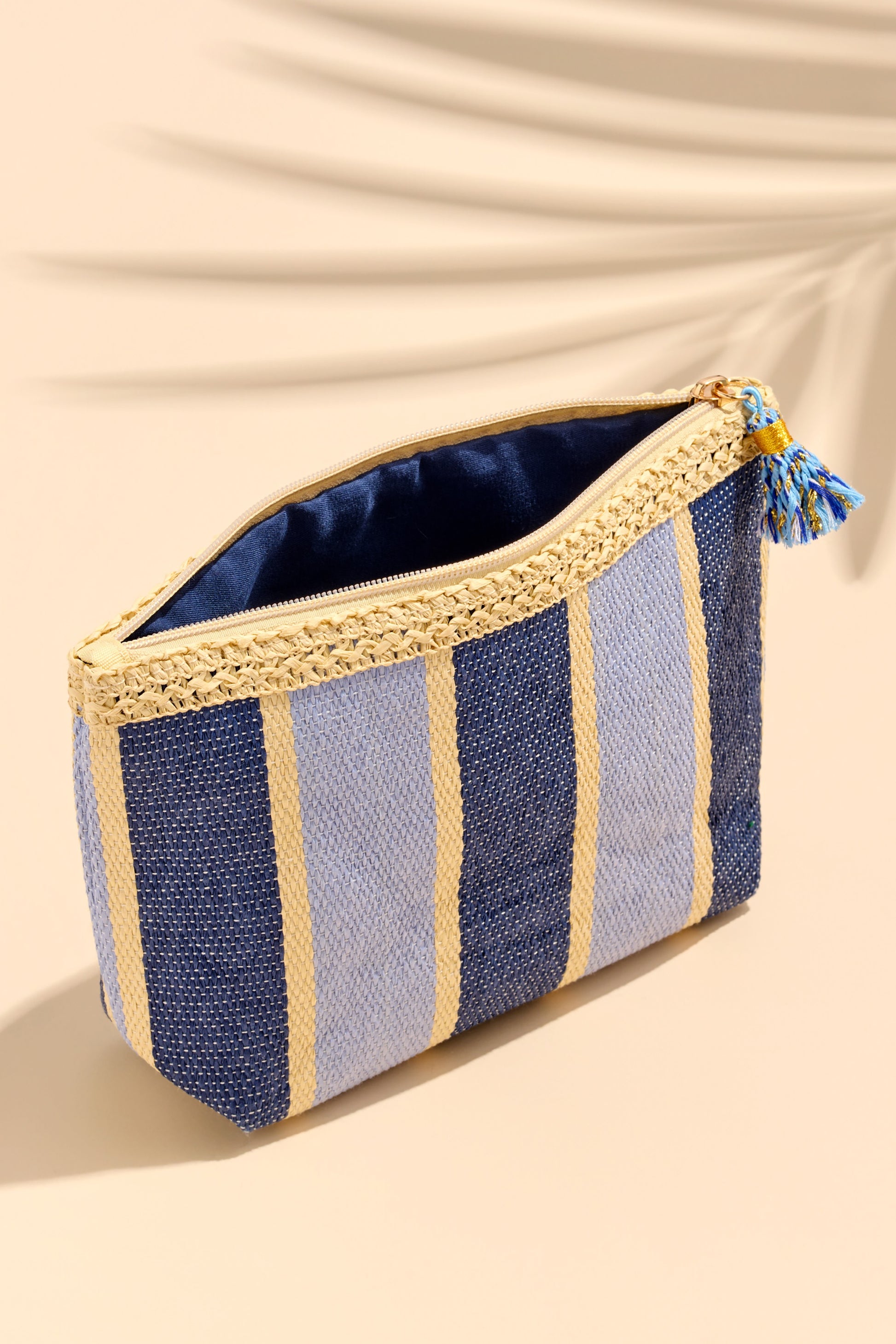 The Capri Zip Pouch by South Moon Under features blue multi stripes, a gold zipper, and a blue tassel, echoing classic beach tote style. Shown against a beige background with a soft palm leaf shadow.