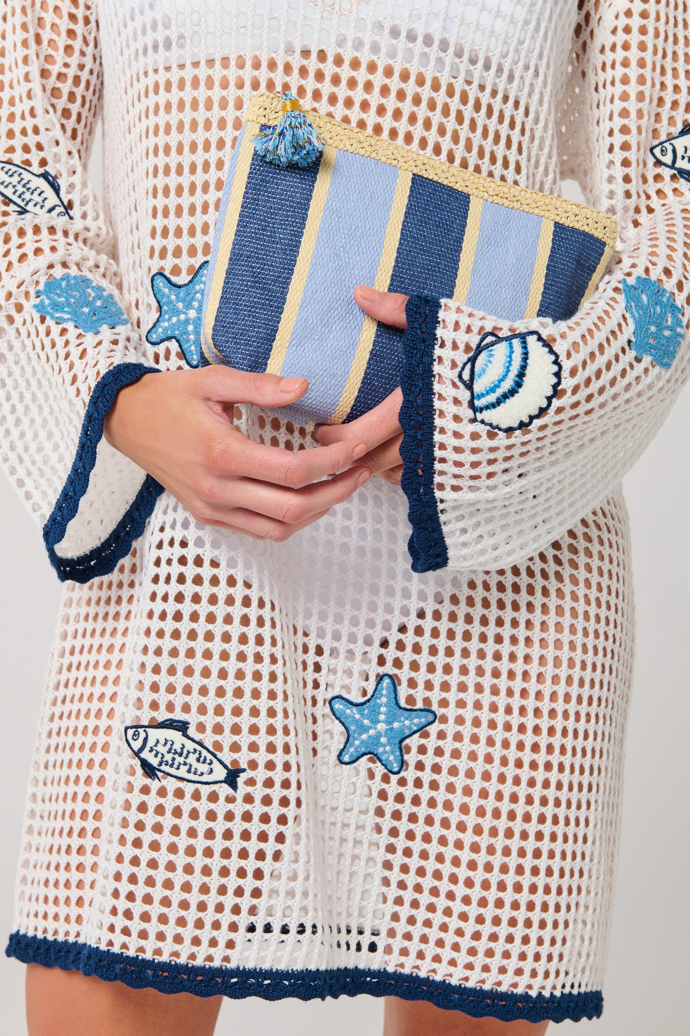 A person in a white mesh dress with blue sea-themed patches holds the Capri Zip Pouch by South Moon Under, featuring blue and yellow stripes and a fun pom-pom detail.
