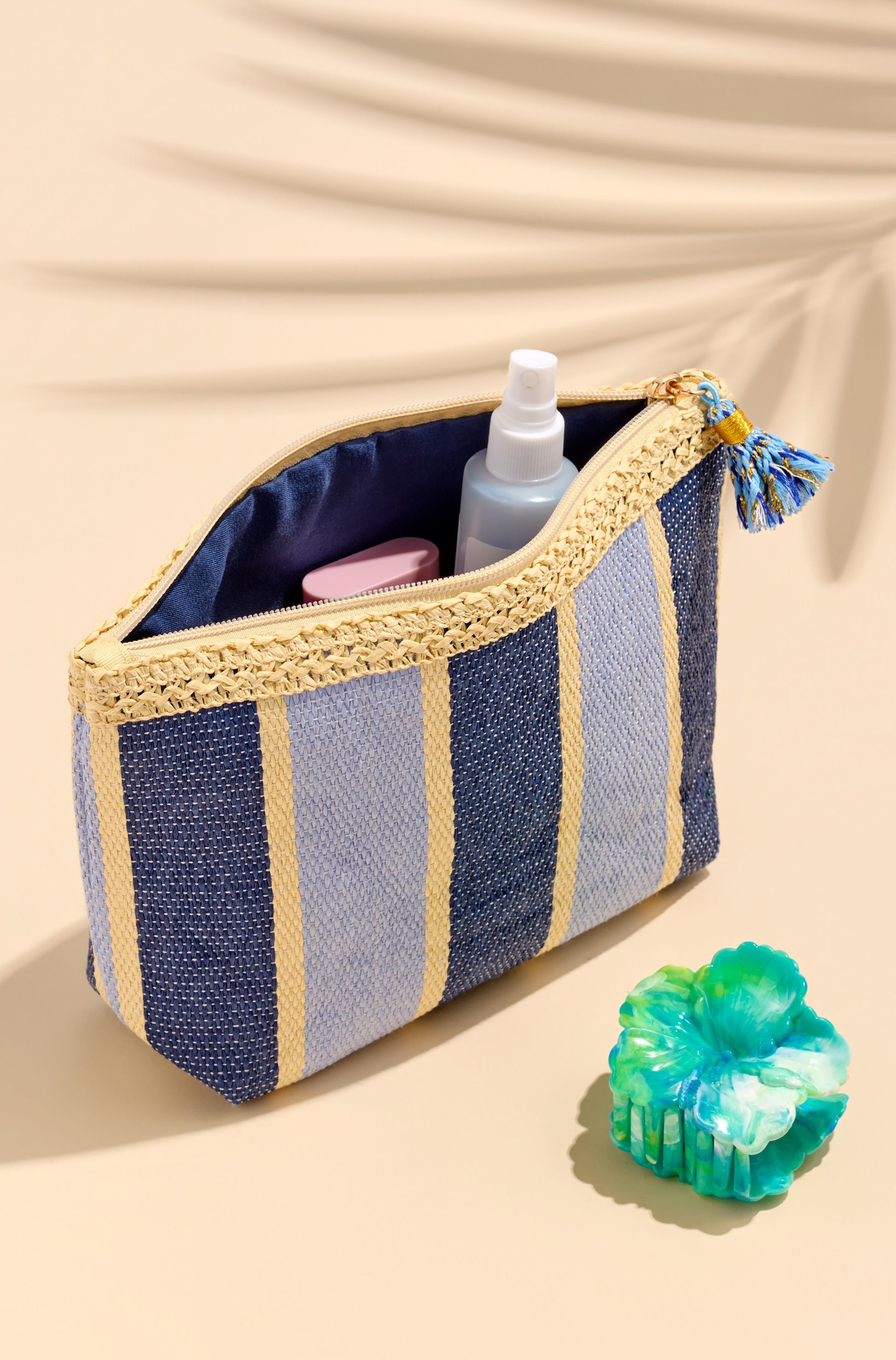 The Capri Zip Pouch by South Moon Under, featuring blue and yellow stripes and a tassel, is unzipped to reveal toiletries. Next to it, a blue and green decorative soap rests on a beige surface with a leaf shadow.