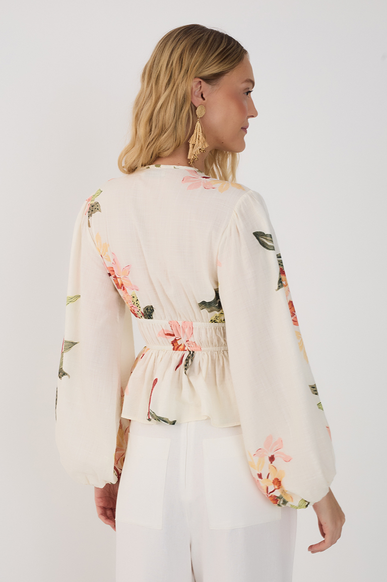A woman with wavy blonde hair and gold earrings models the Dress To Flourish Tie Front Blouse, a cream bohemian top with botanical print, paired with white pants. She stands facing away from the camera against a plain background.
