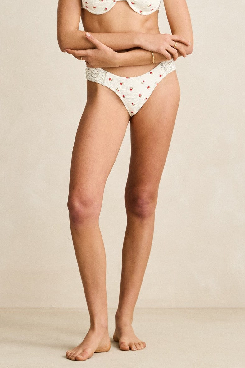 A woman stands barefoot against a plain backdrop, wearing Rhythm's Cream Mila Soft Side Holiday Pant—a mid-rise, cream panty with moderate coverage and a small red floral print. Her face is not shown.