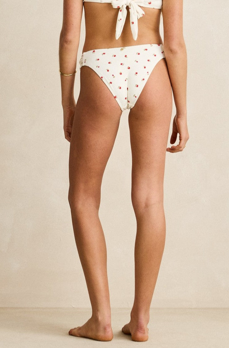 A woman stands barefoot on a light floor, shown from the back, wearing the Rhythm Cream Mila Soft Side Holiday Pant with moderate coverage and mid-rise waist, featuring a small red cherry print and a matching white tie-back bikini top.