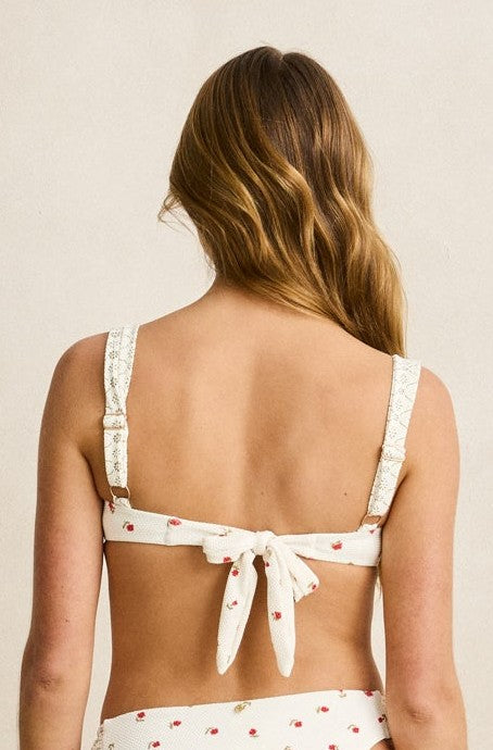 A woman with wavy light brown hair is shown from the back wearing the Rhythm Cream Mila Soft Strap Balconette Top, featuring a white base with a small red floral print, lace trim, and adjustable straps. The background is plain and neutral.