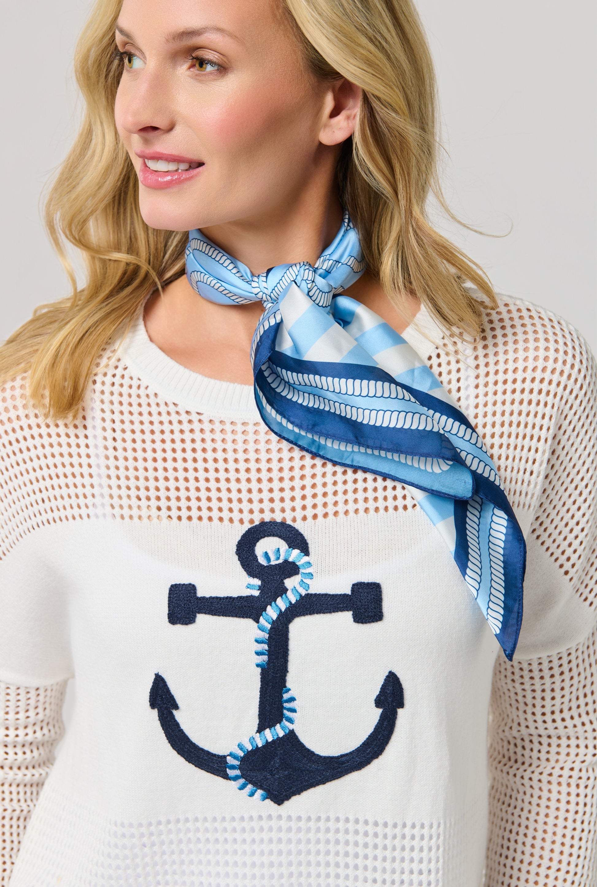A woman smiles and looks to the side, wearing a white mesh sweater with a navy anchor design and the Coastal Scarf by South Moon Under tied sailor-chic around her neck.