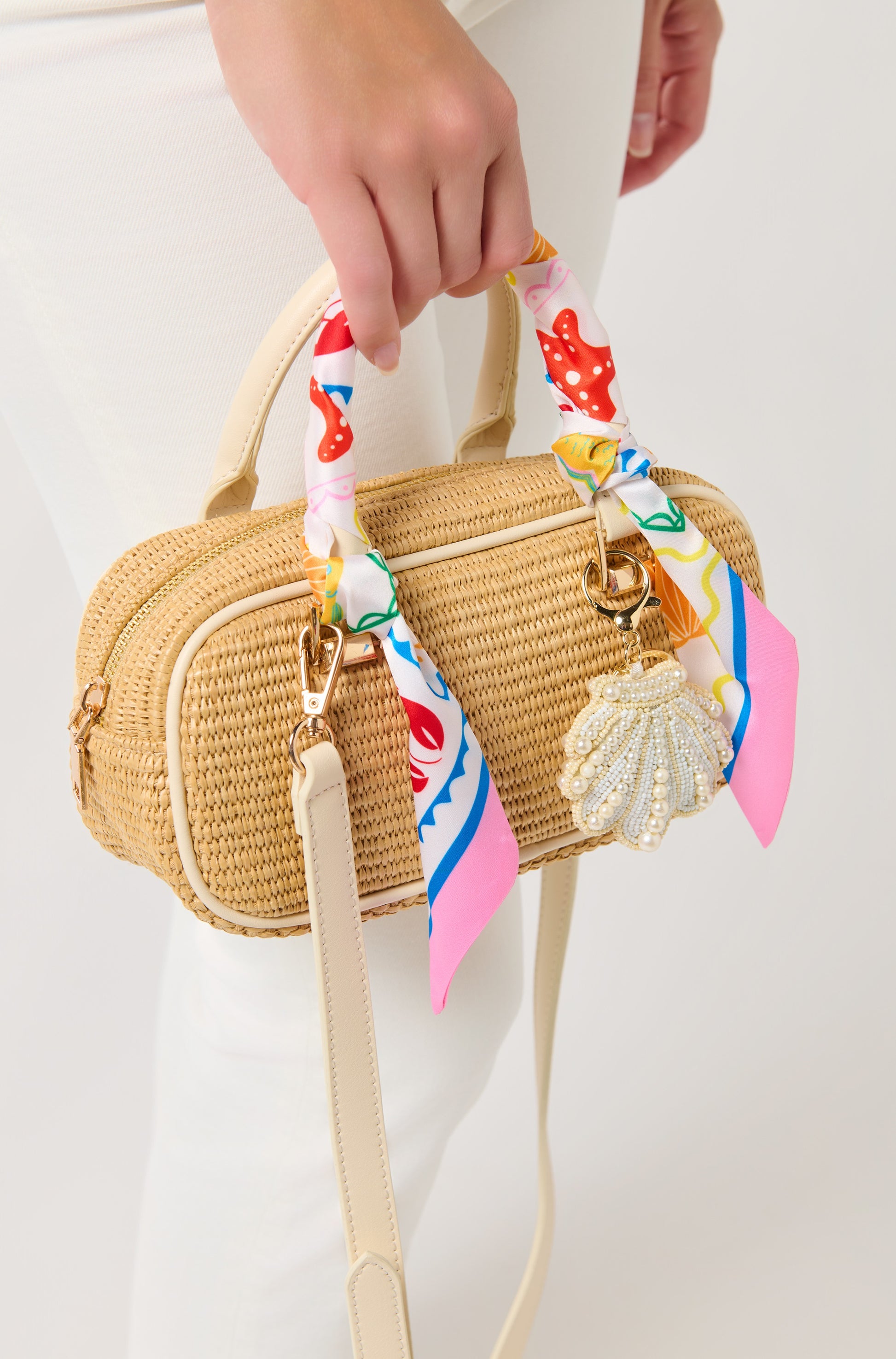 A person in white pants holds a small straw handbag accessorized with South Moon Under’s Assorted Set of 2 Tropical Lobster Twilly Scarves wrapped on the handles and a beaded seashell charm attached.