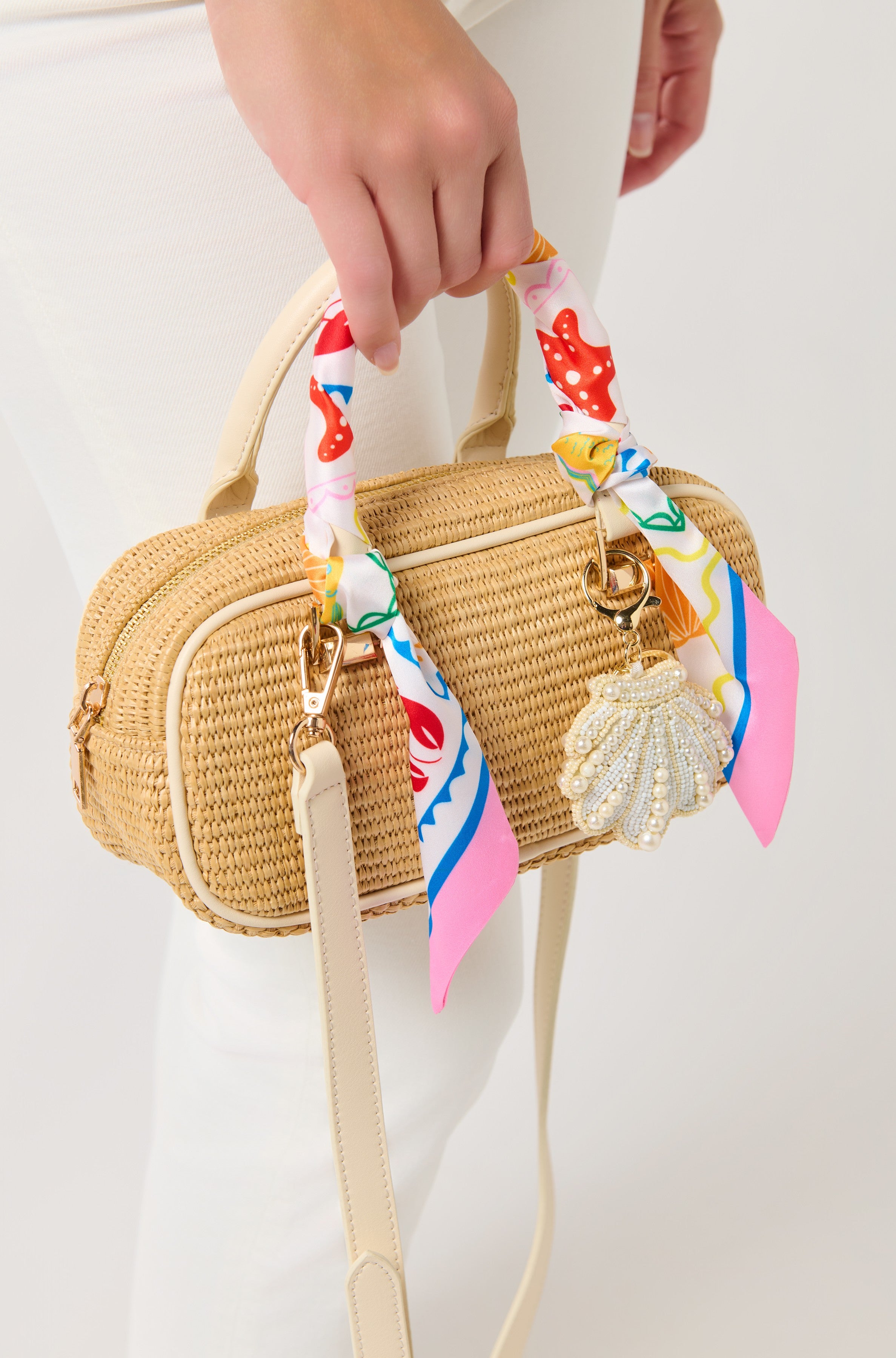 A person in white pants holds a small straw handbag accessorized with South Moon Under’s Assorted Set of 2 Tropical Lobster Twilly Scarves wrapped on the handles and a beaded seashell charm attached.