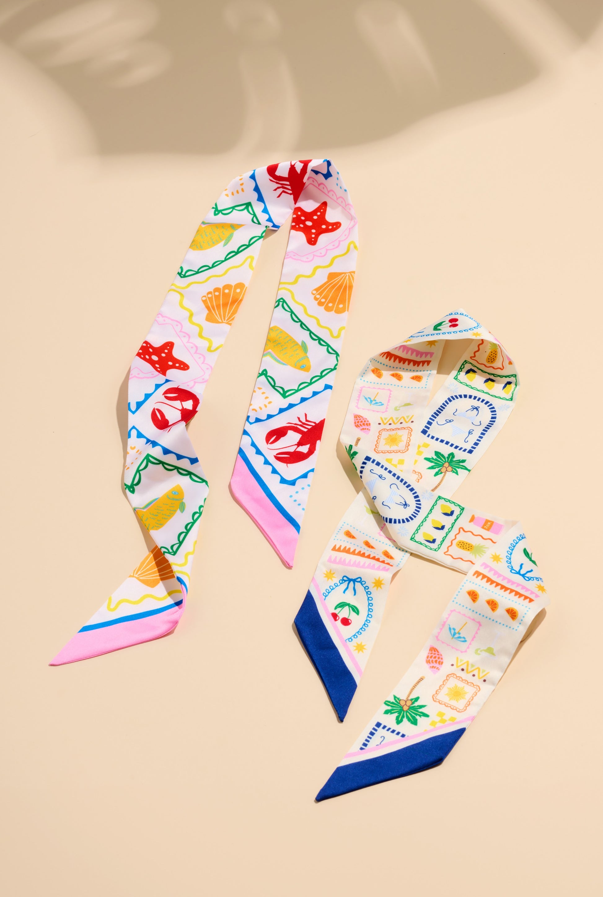 The South Moon Under Assorted Set of 2 Tropical Lobster Twilly Scarves in White Multi displays vibrant, beachy motifs and pointed ends, laid side by side on a beige background. Each scarf is crafted from polyester silk.